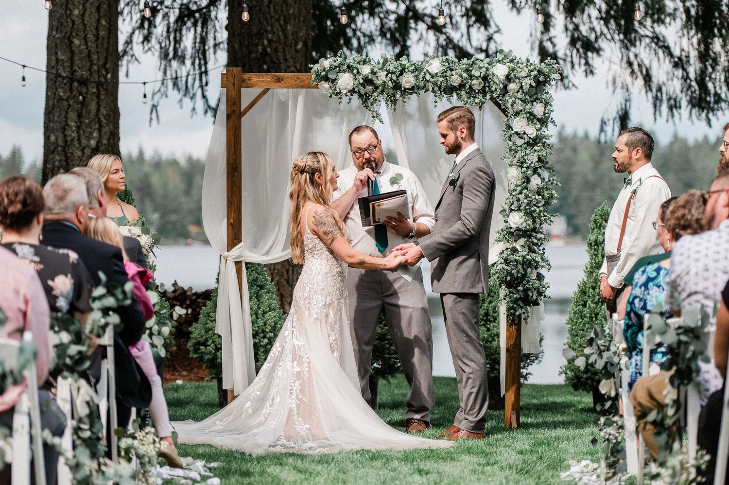 Outdoor wedding ceremony by a lake with bride in lace gown, groom in gray suit, and officiant under floral arch.