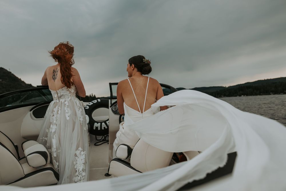 Two brides in white gowns stand on a boat with veils flowing in the wind over a scenic lake.