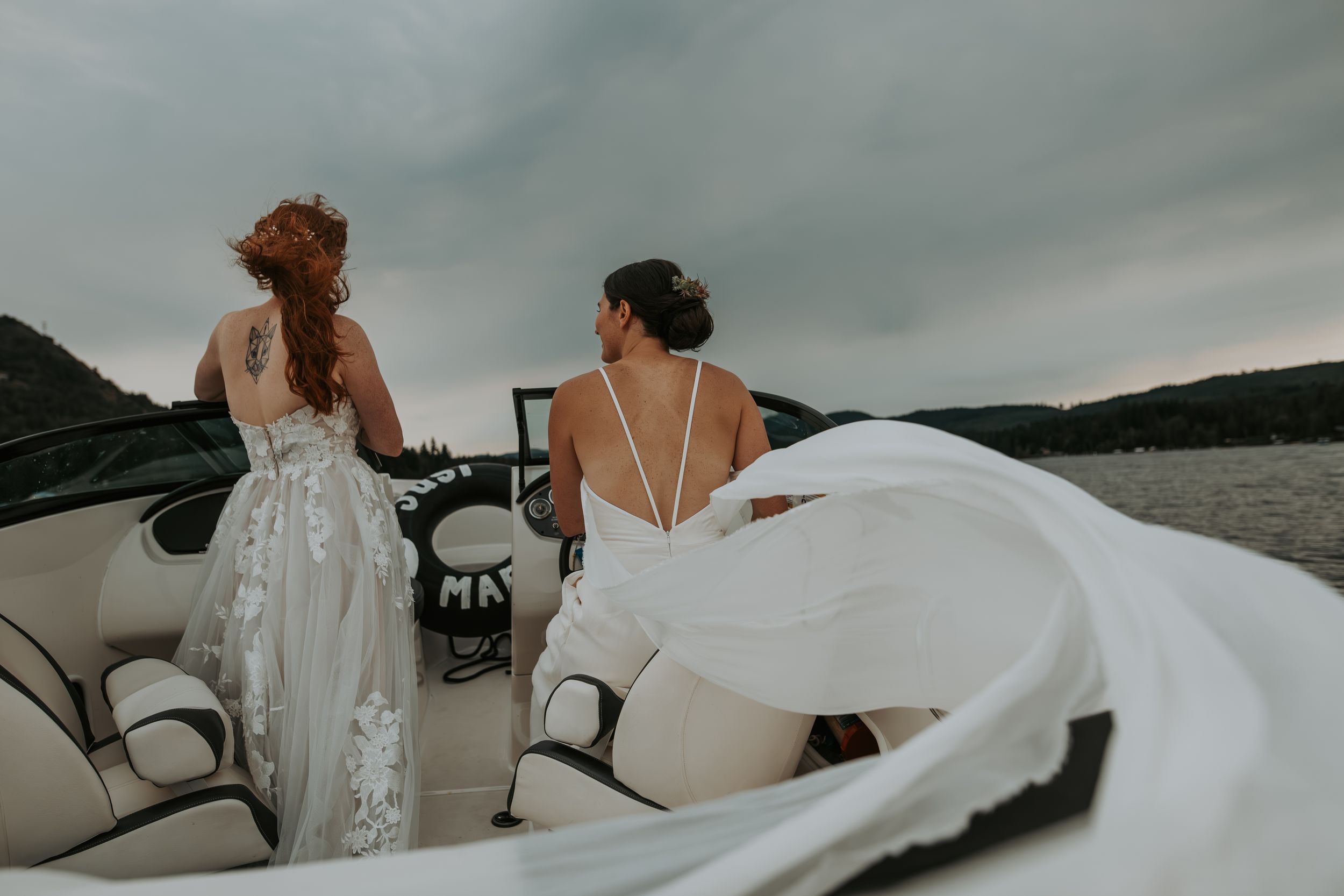 Two brides in white gowns stand on a boat with veils flowing in the wind over a scenic lake.