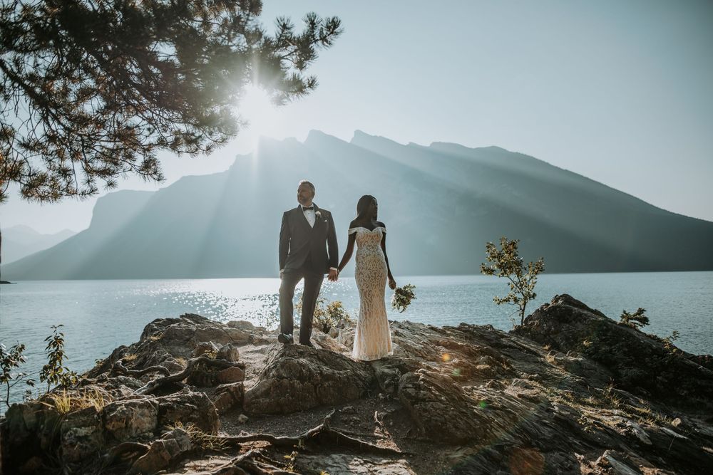Bride and groom stand on rocky lakeside with stunning mountain backdrop and sunburst at their outdoor wedding.