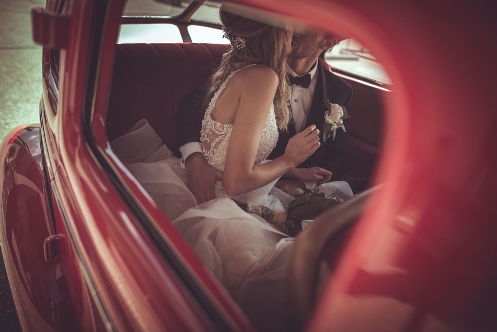 Bride and groom sharing intimate moment inside vintage red classic car on wedding day.