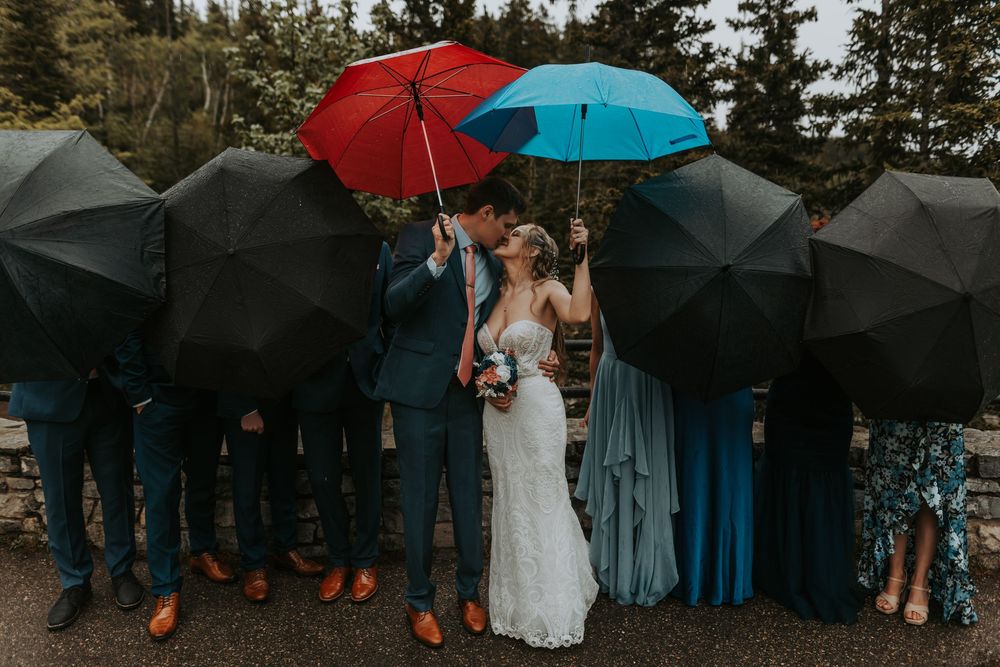 Wedding party shielding couple with colorful umbrellas as bride and groom kiss in the rain.