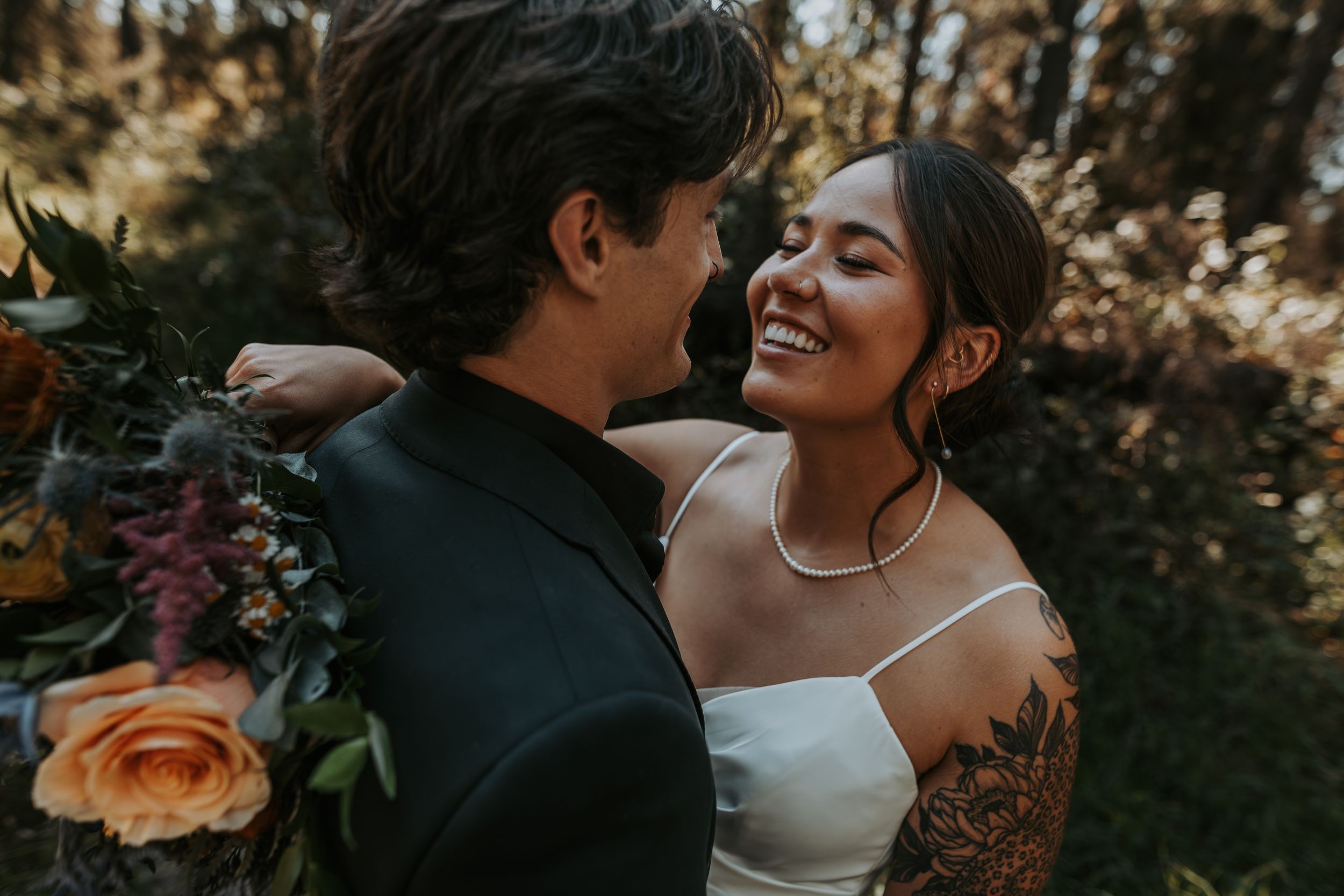 Smiling tattooed bride in white dress embracing groom while holding colorful wedding bouquet in forest.