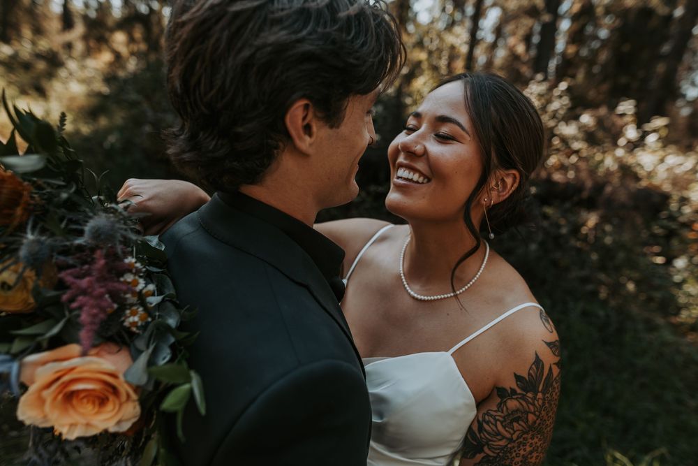 Smiling tattooed bride in white dress embracing groom while holding colorful wedding bouquet in forest.