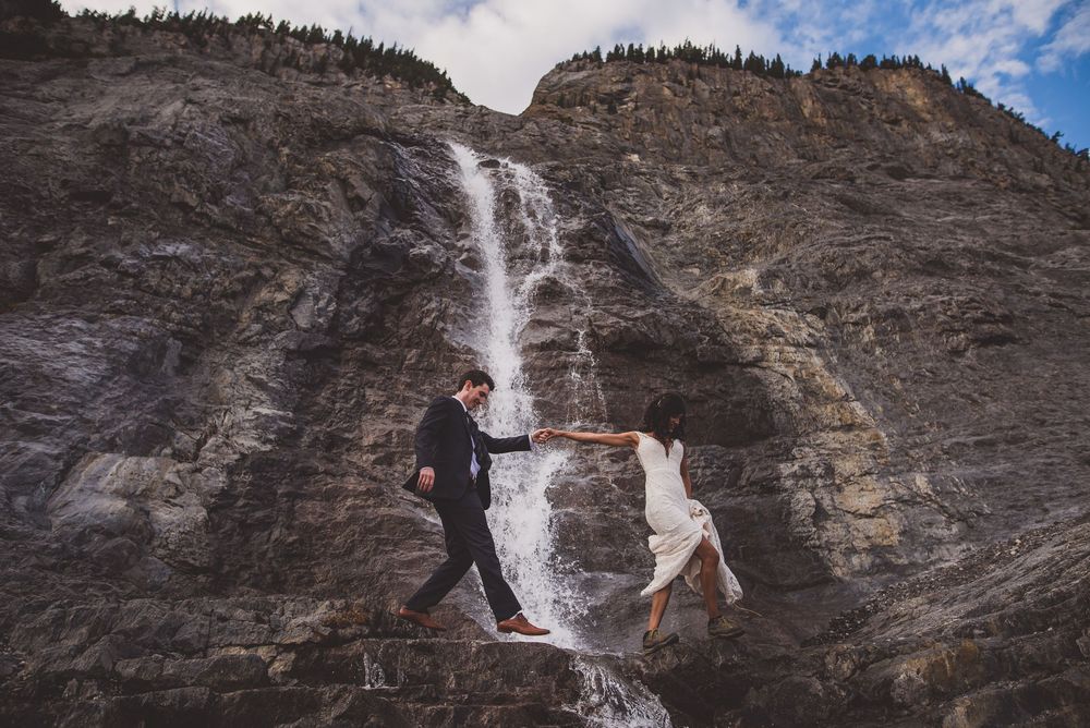Bride and groom hold hands while climbing rocks beside a stunning cascading waterfall in the mountains.