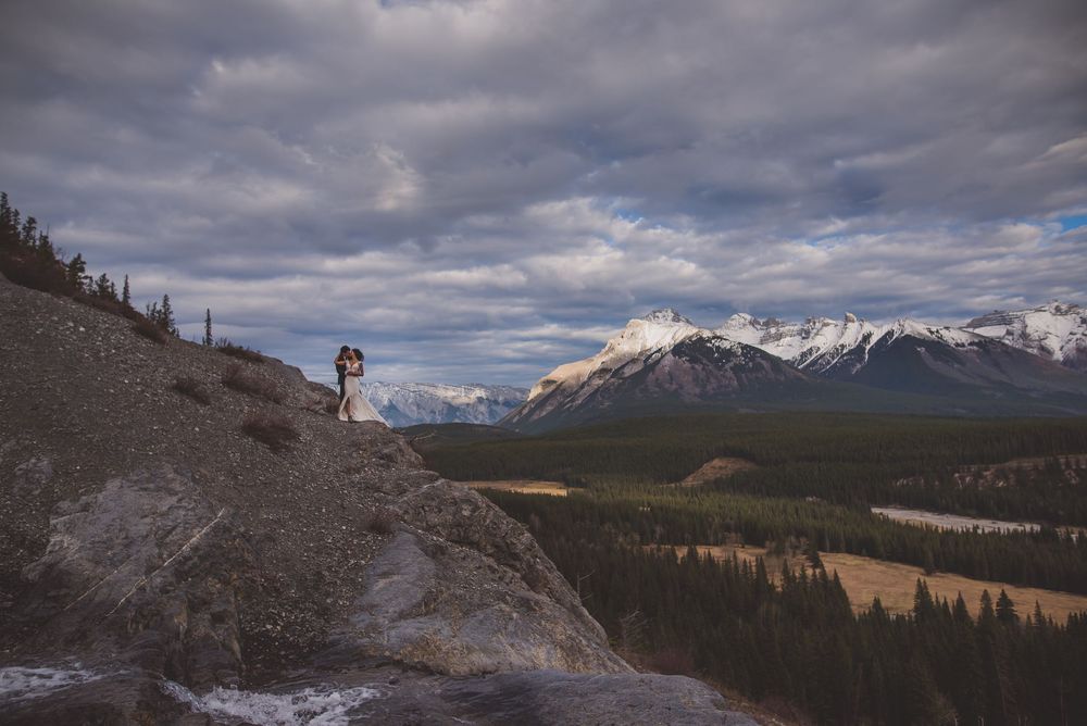 Bride and groom stand on a rocky ridge overlooking a vast mountain valley with snow-capped peaks.
