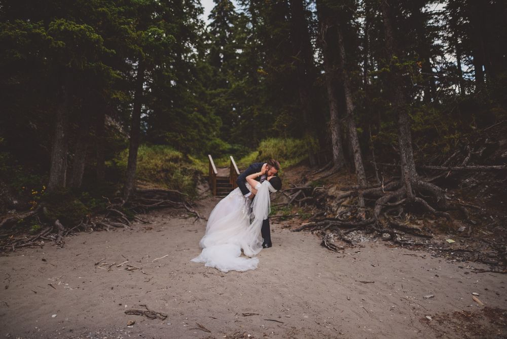 Groom dips bride in a romantic kiss on a sandy forest beach surrounded by tall evergreen trees.