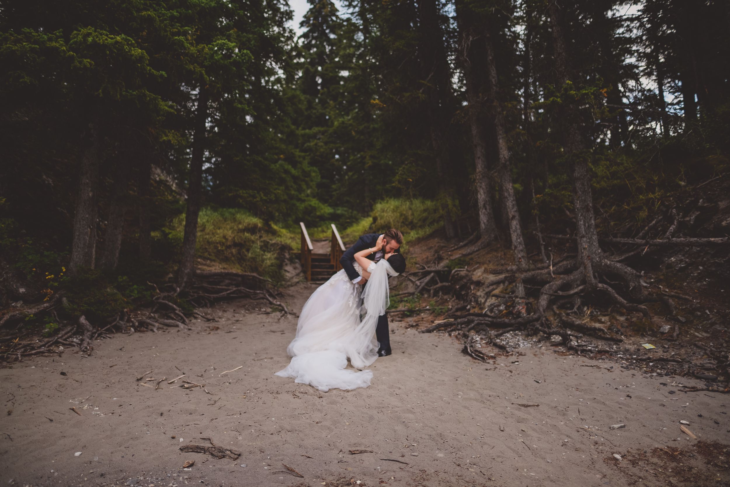 Groom dips bride in a romantic kiss on a sandy forest beach surrounded by tall evergreen trees.