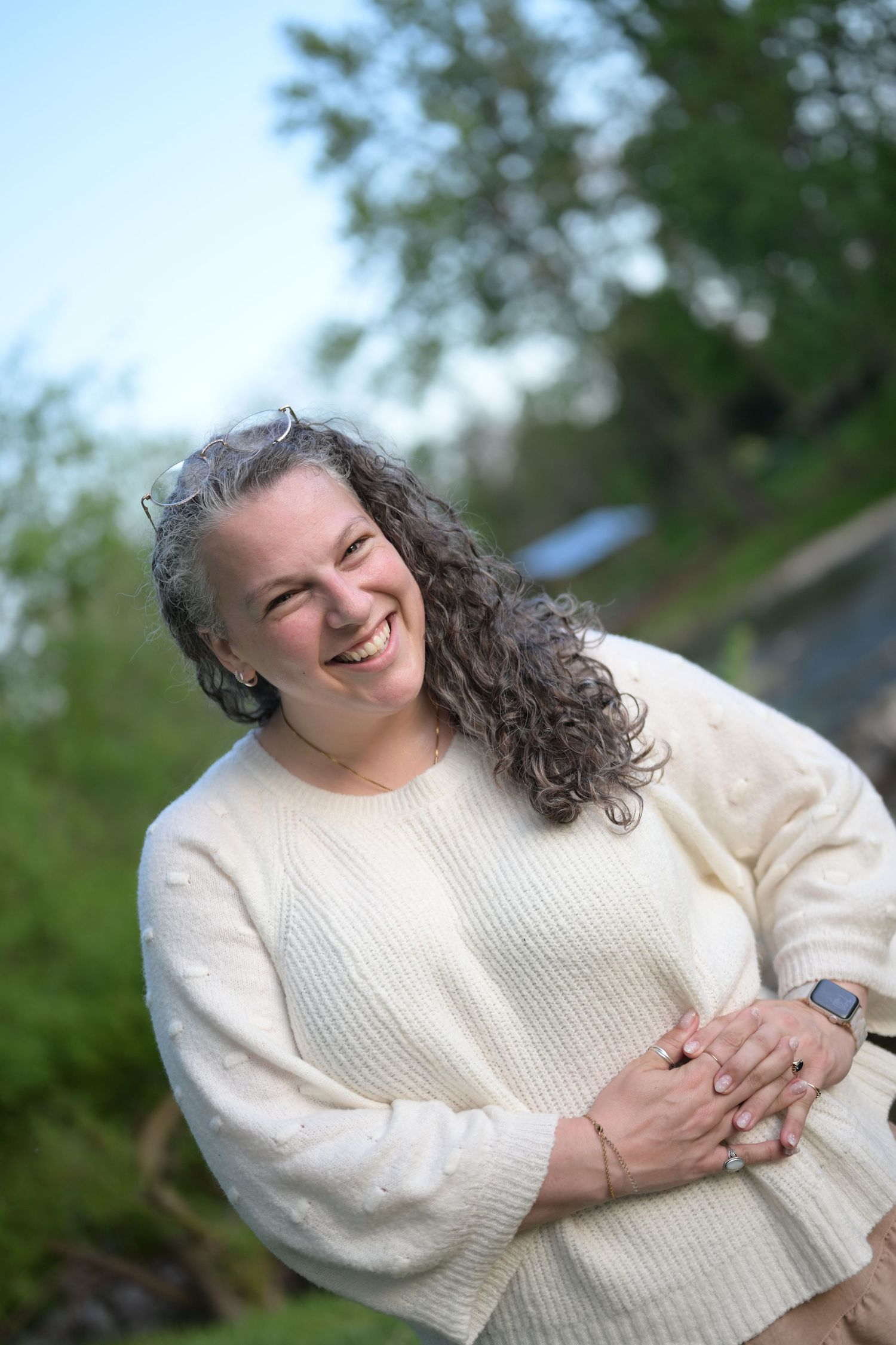 Middle-aged woman in white tank top smiling outdoors in a sunny park setting with green trees in background.