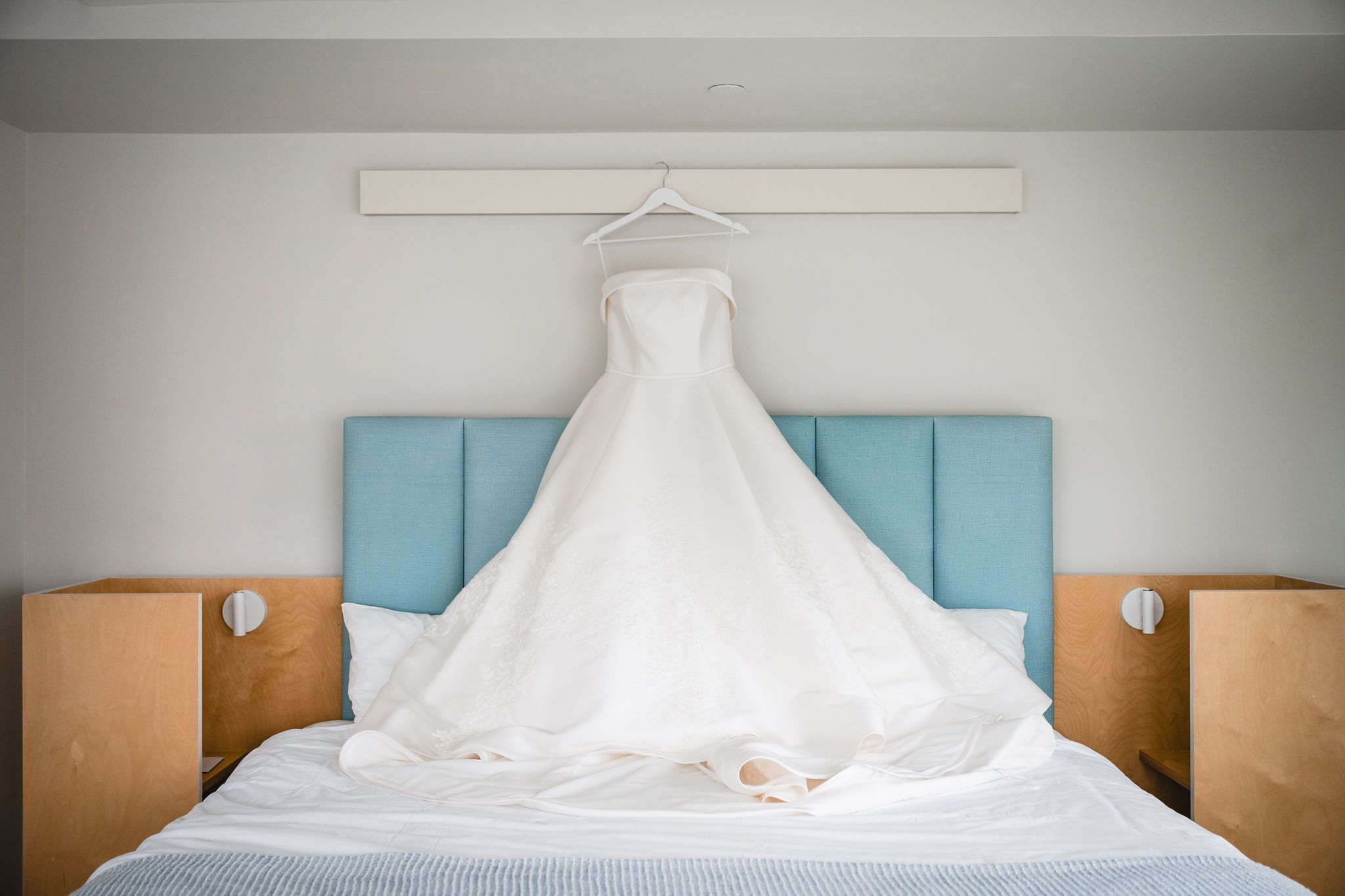 White wedding gown hanging above a bed with a blue headboard in a bright hotel room.