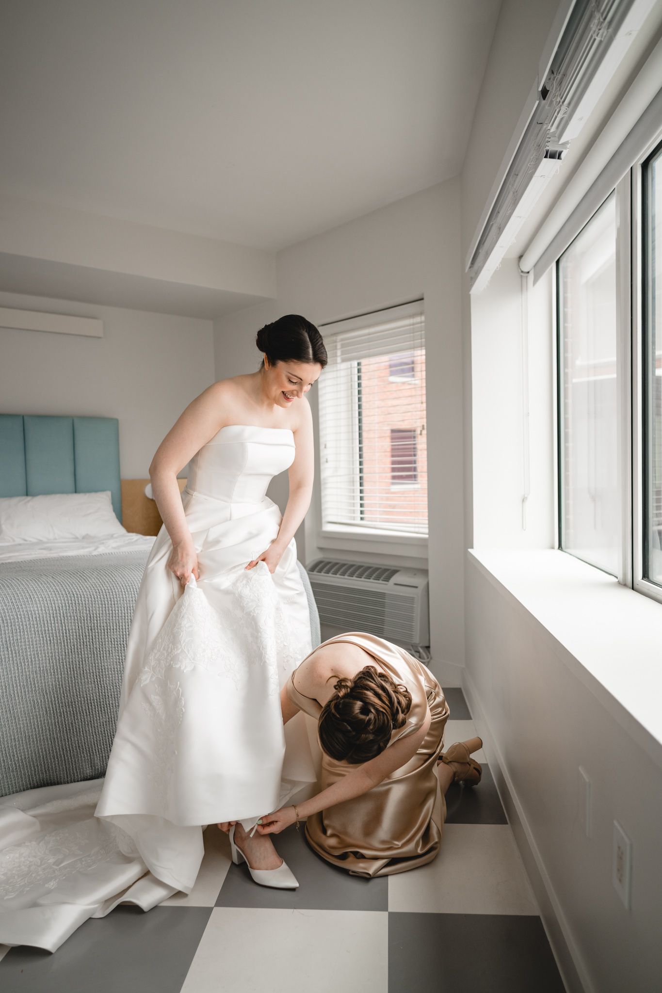 Bride in white strapless gown getting shoes fastened by bridesmaid in gold dress in bright hotel room.