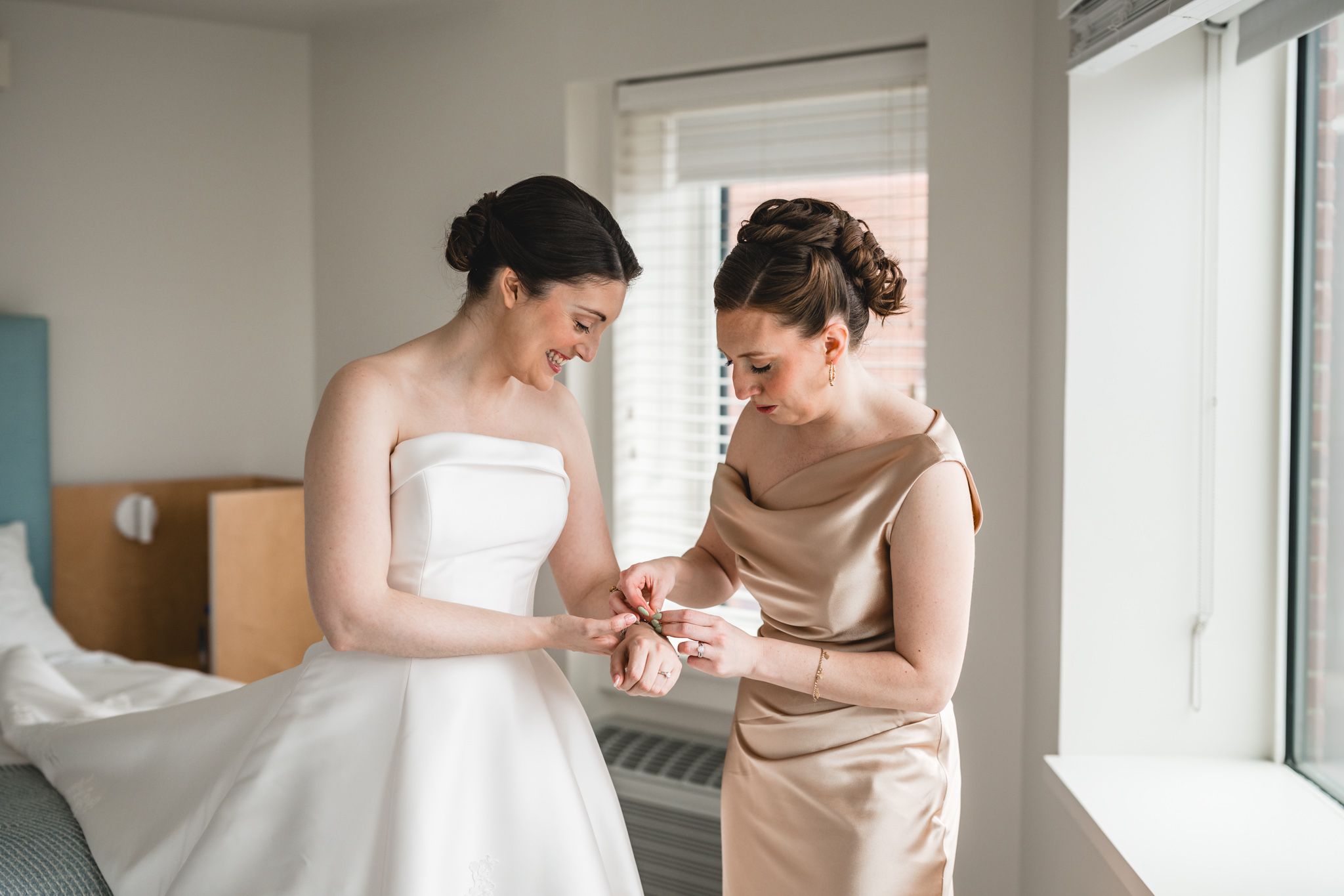 Bride in white strapless gown smiling as bridesmaid in gold dress helps her with jewelry near a bright window.
