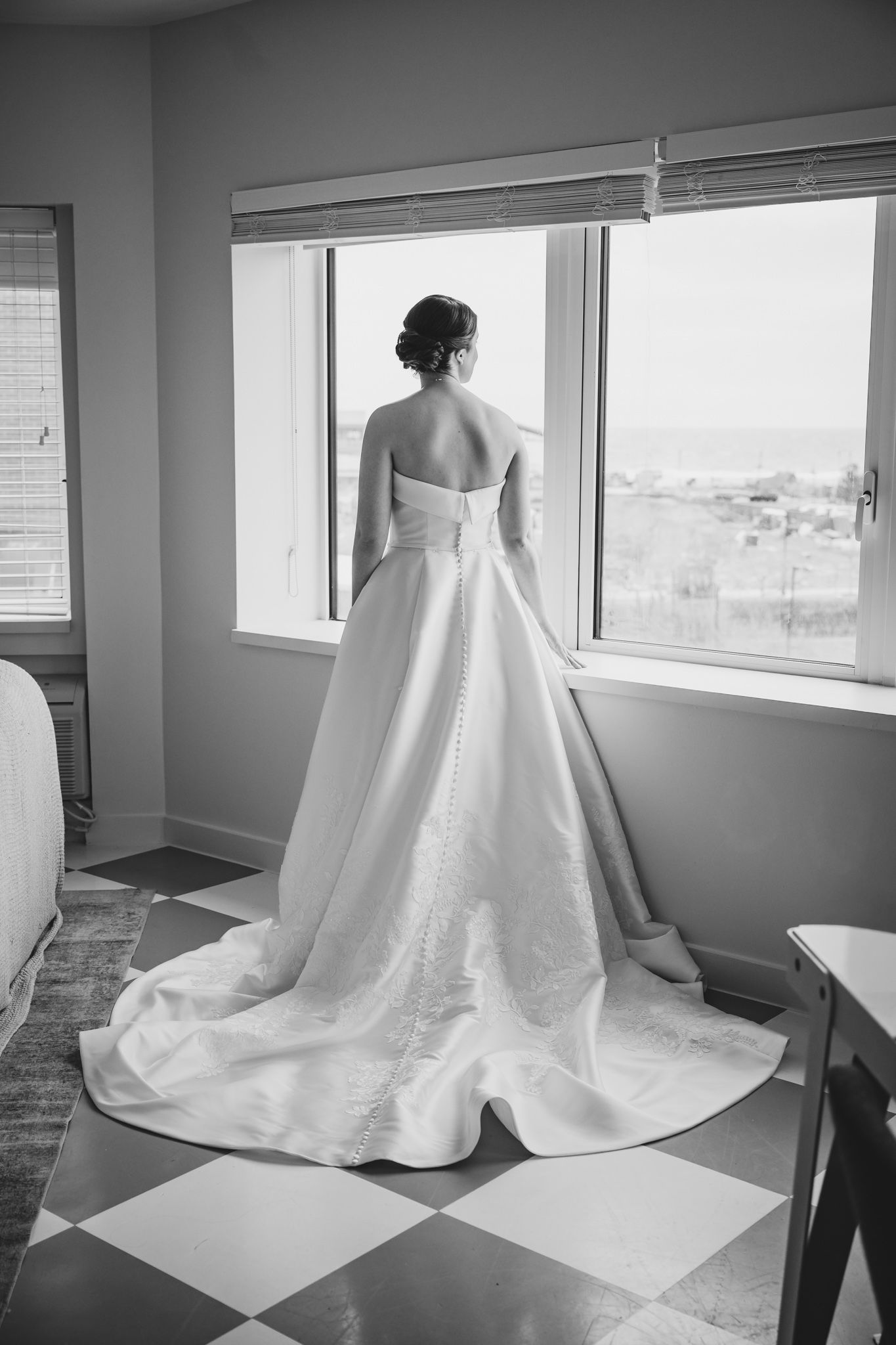 Black and white photo of bride in strapless ball gown with long train gazing out window before wedding.