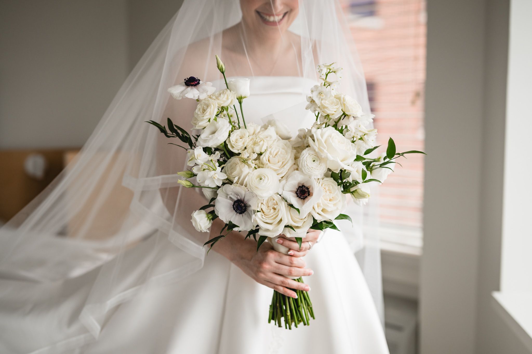 Bride holding elegant white bouquet with anemones and roses, wearing a flowing veil and strapless wedding gown.