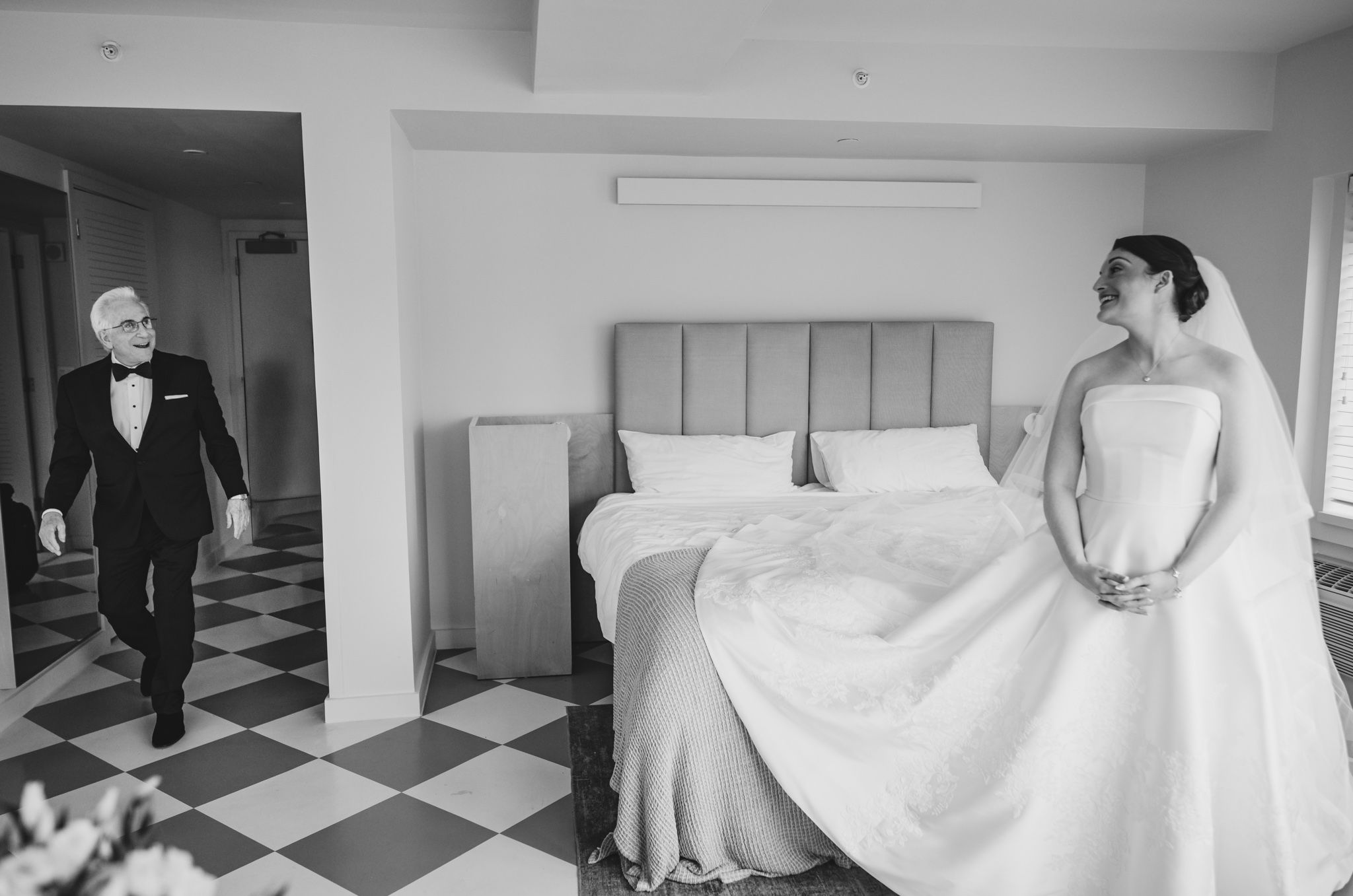 Black and white photo of groom in tuxedo approaching bride in wedding dress in hotel room.