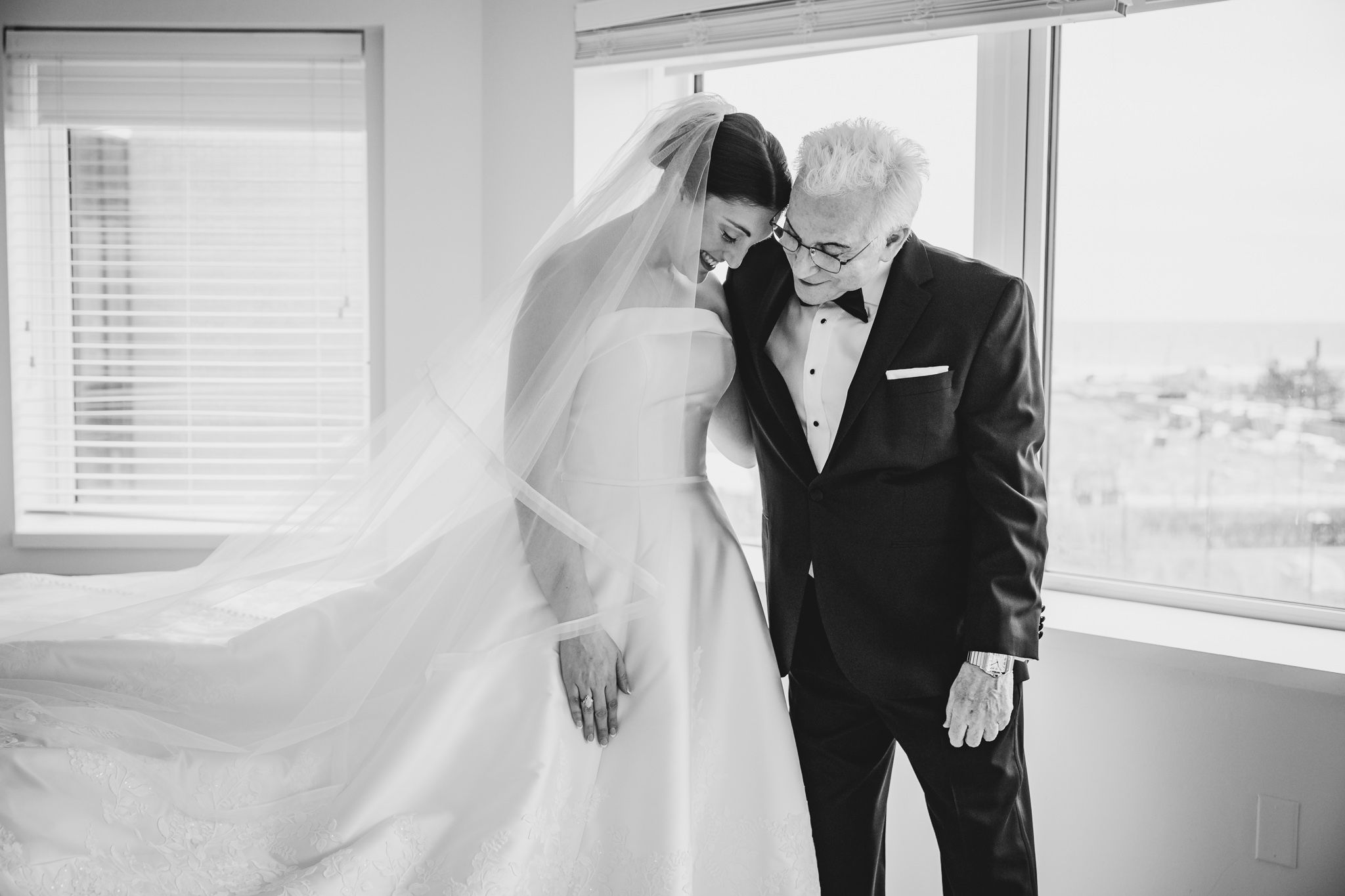 Bride and older gentleman sharing tender moment in black and white photo, with veil flowing dramatically.