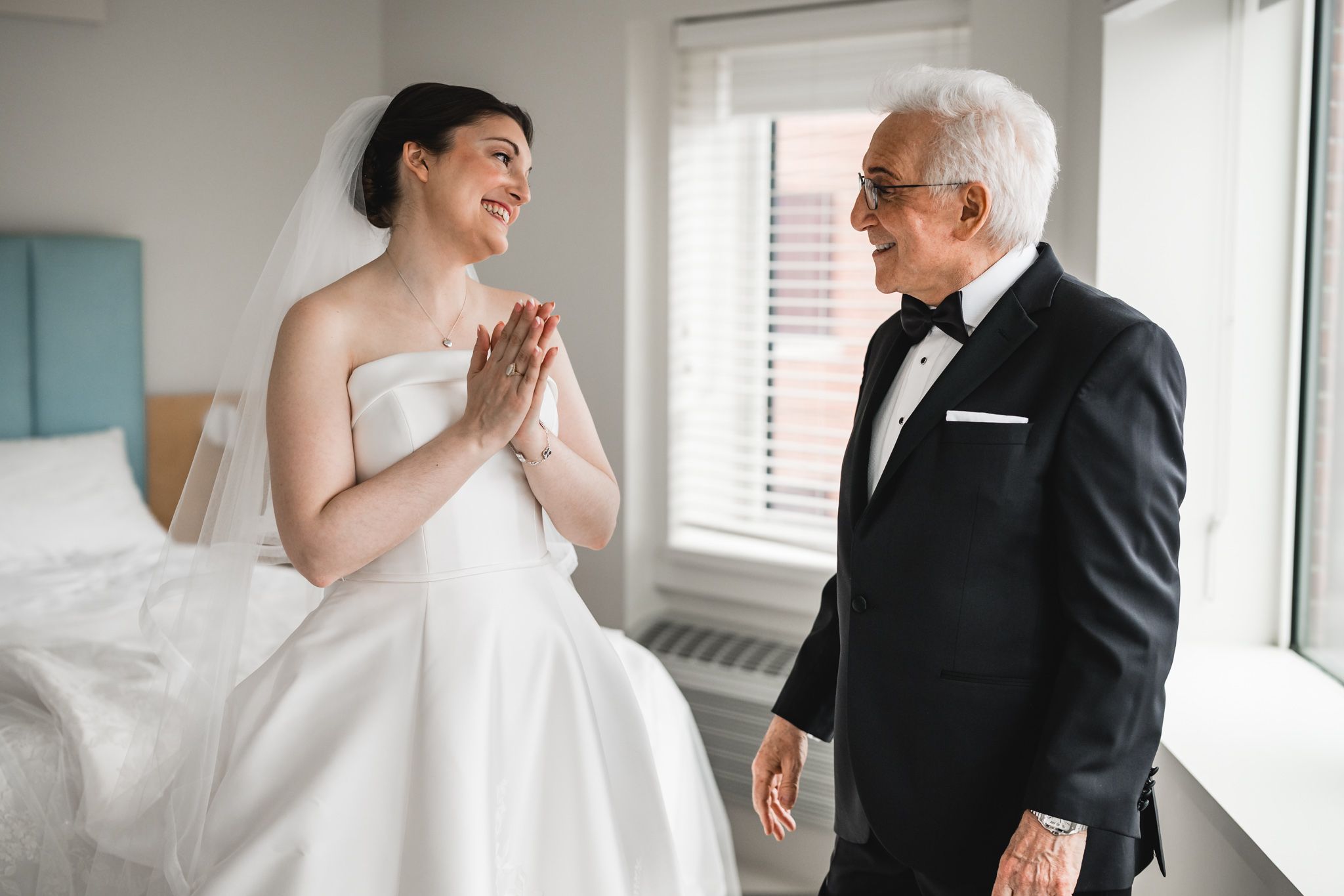 Bride in white gown smiles at older man in tuxedo during wedding day getting ready moment.