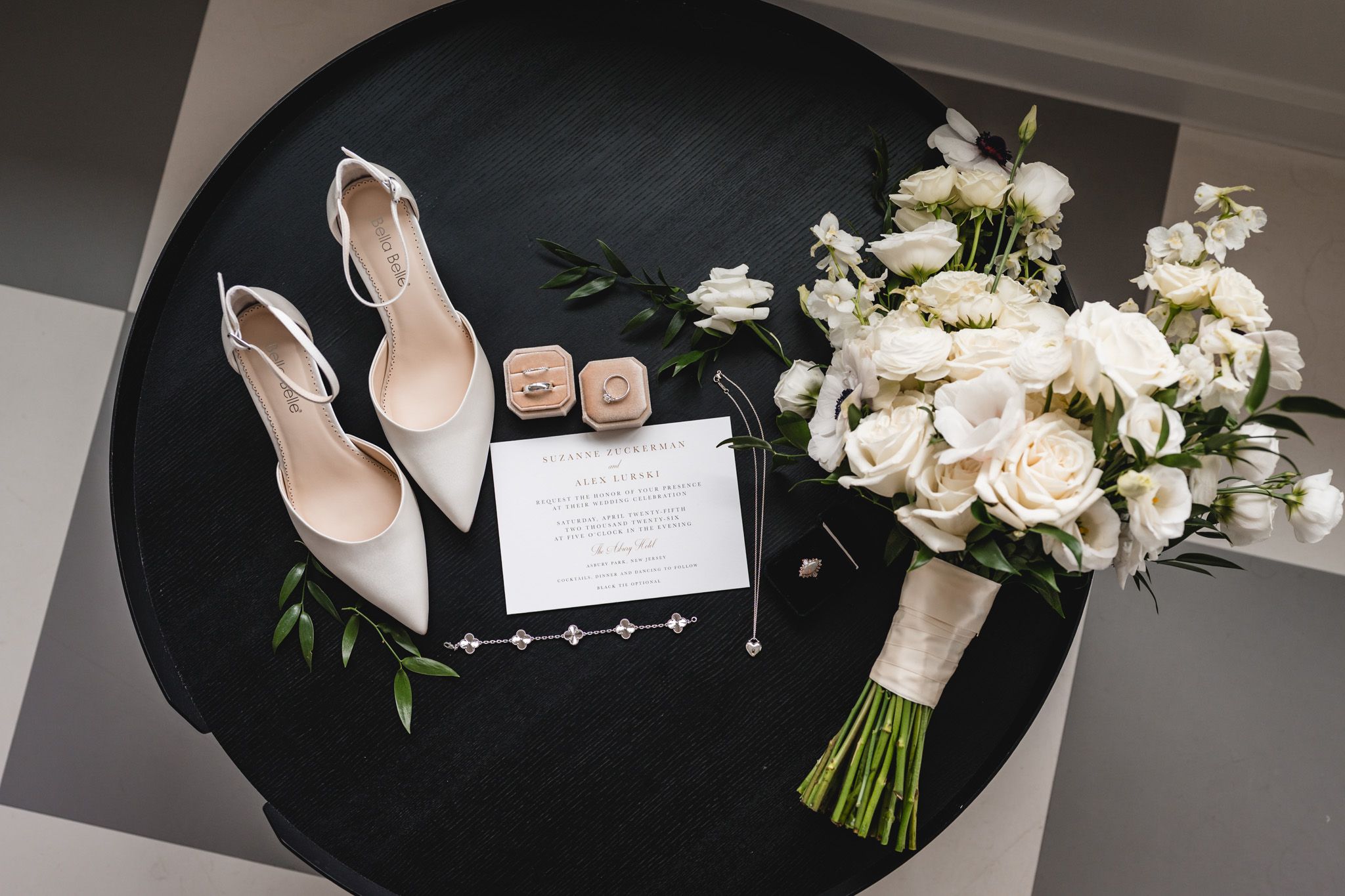 Bridal flat lay with white heels, bouquet, rings, and invitation on a dark round table.