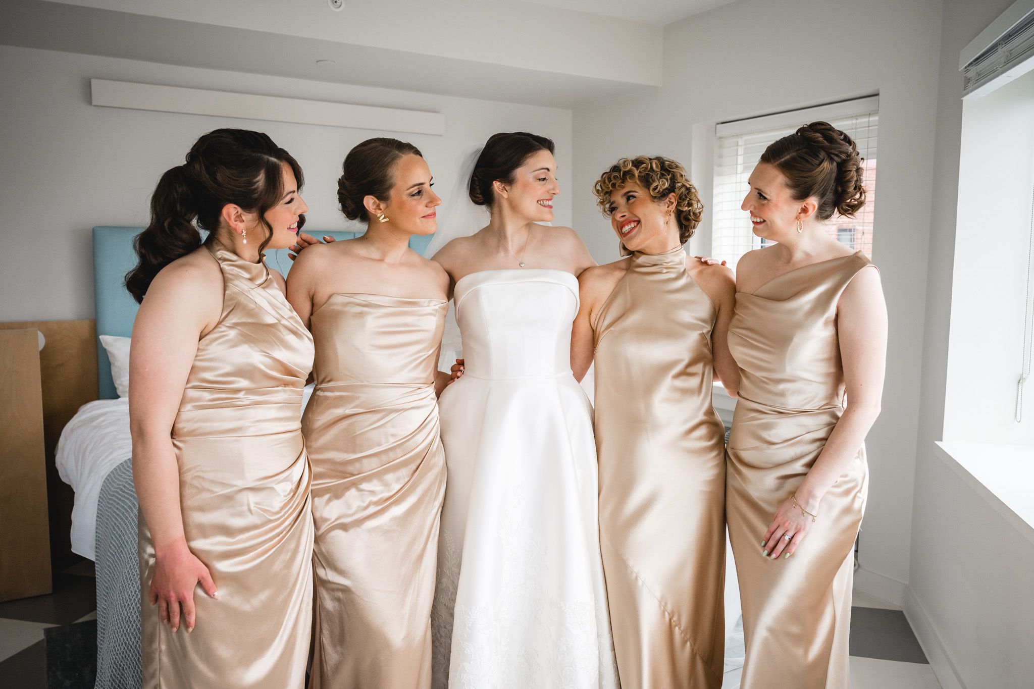 Bride in white gown poses with four bridesmaids in gold satin dresses, smiling together in a bright room.
