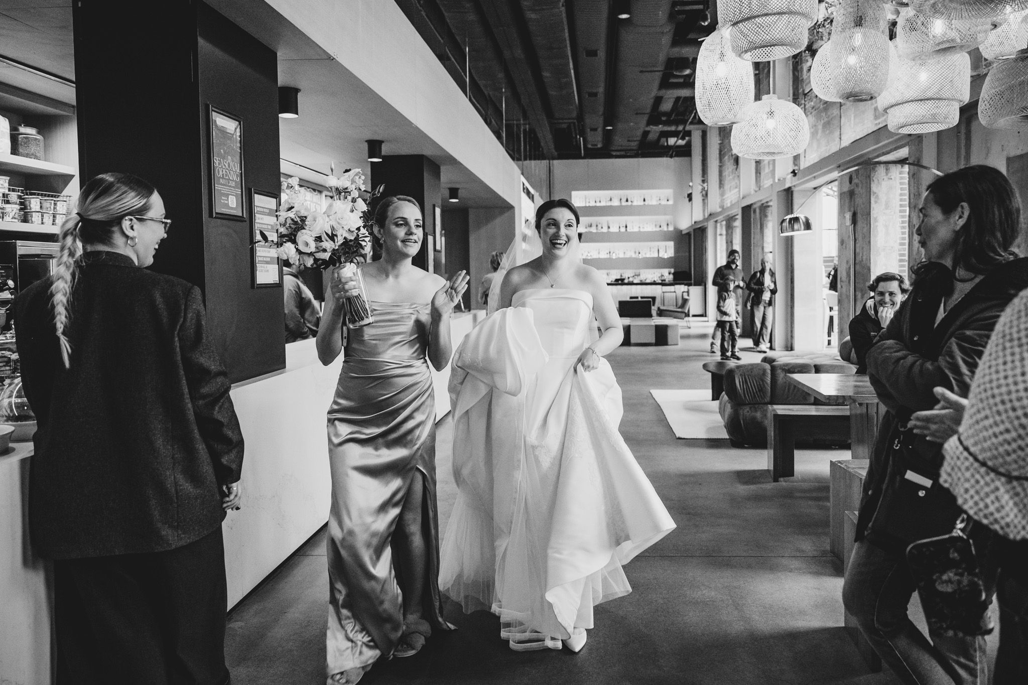 Black and white photo of bride in white gown walking through modern venue with bridesmaids before ceremony.