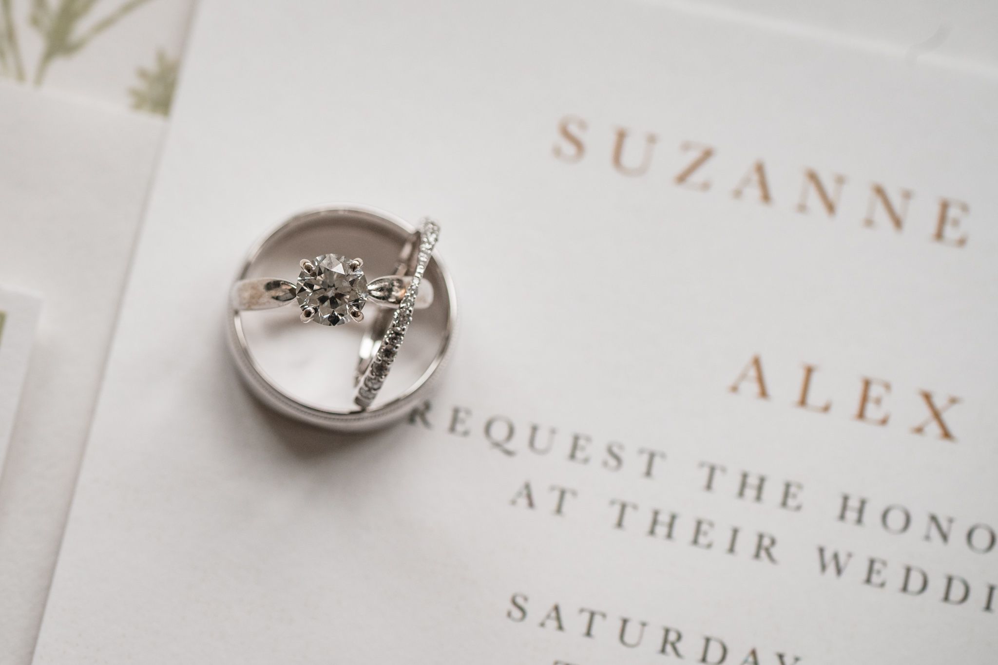 Silver diamond engagement ring resting on a ceramic dish atop elegant wedding invitation stationery.