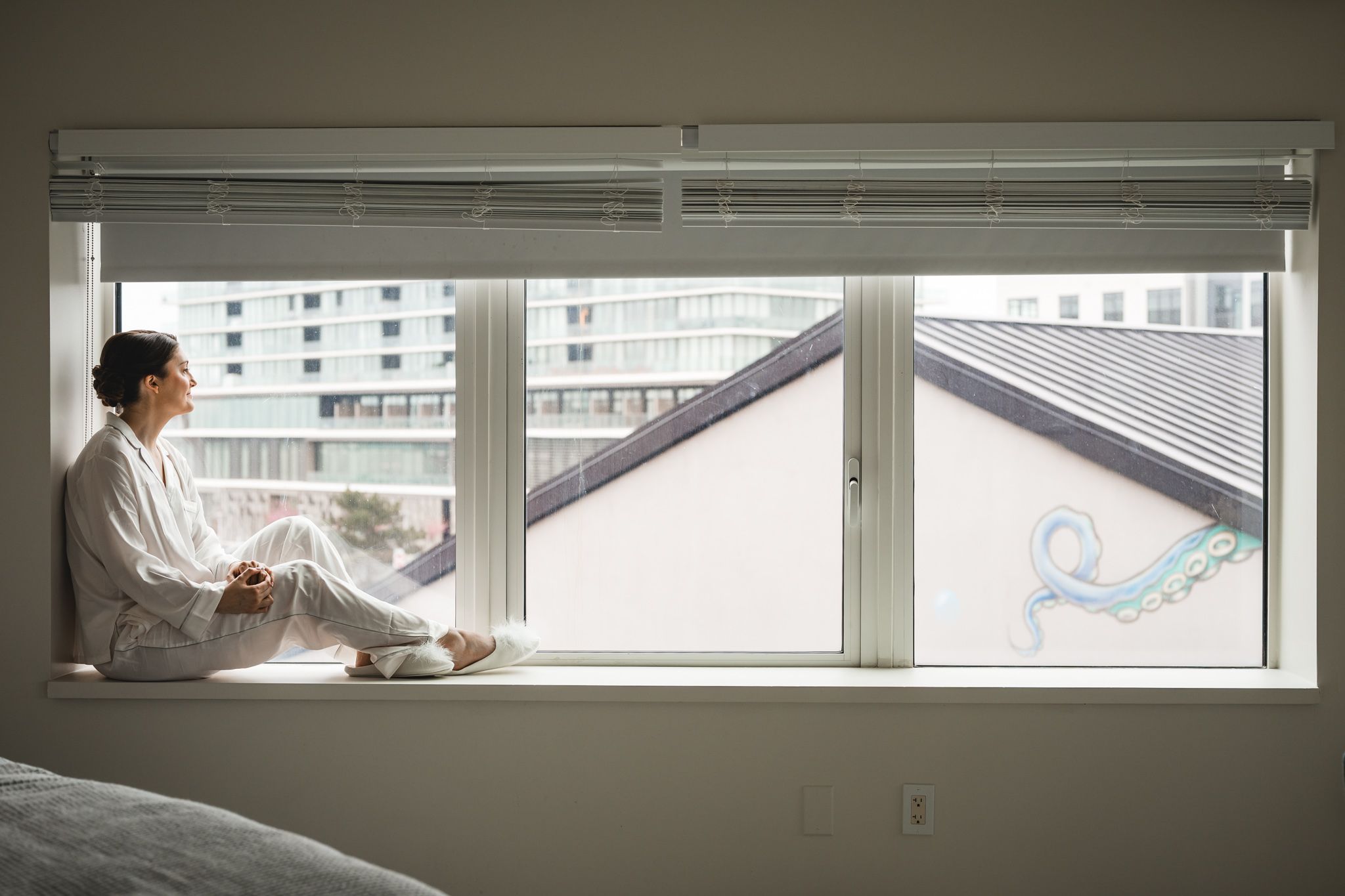 Person in white robe sitting on hotel room windowsill gazing at city skyline.