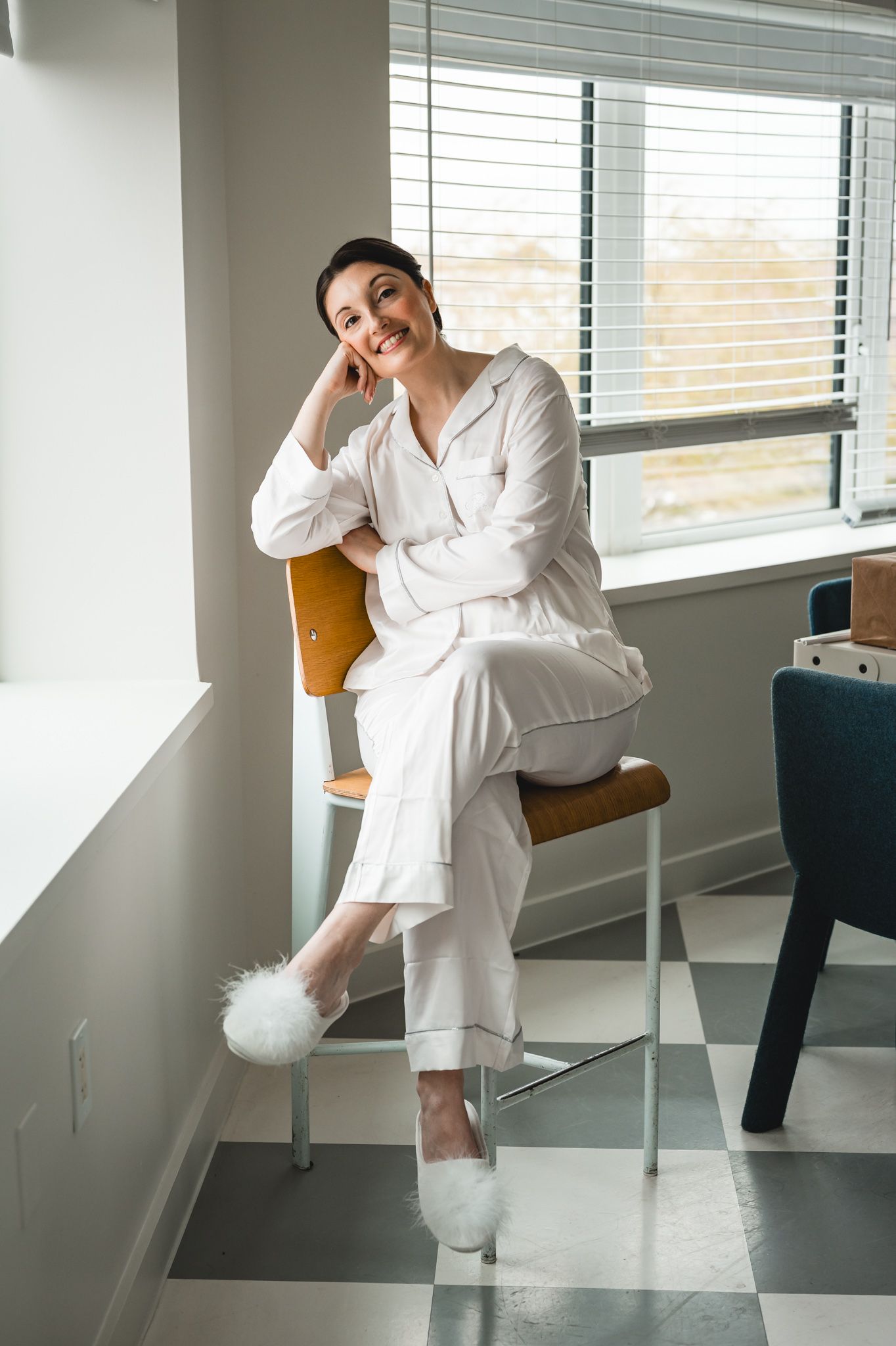 Woman in white pajamas and fluffy slippers sitting on a wooden chair smiling by a window with blinds.