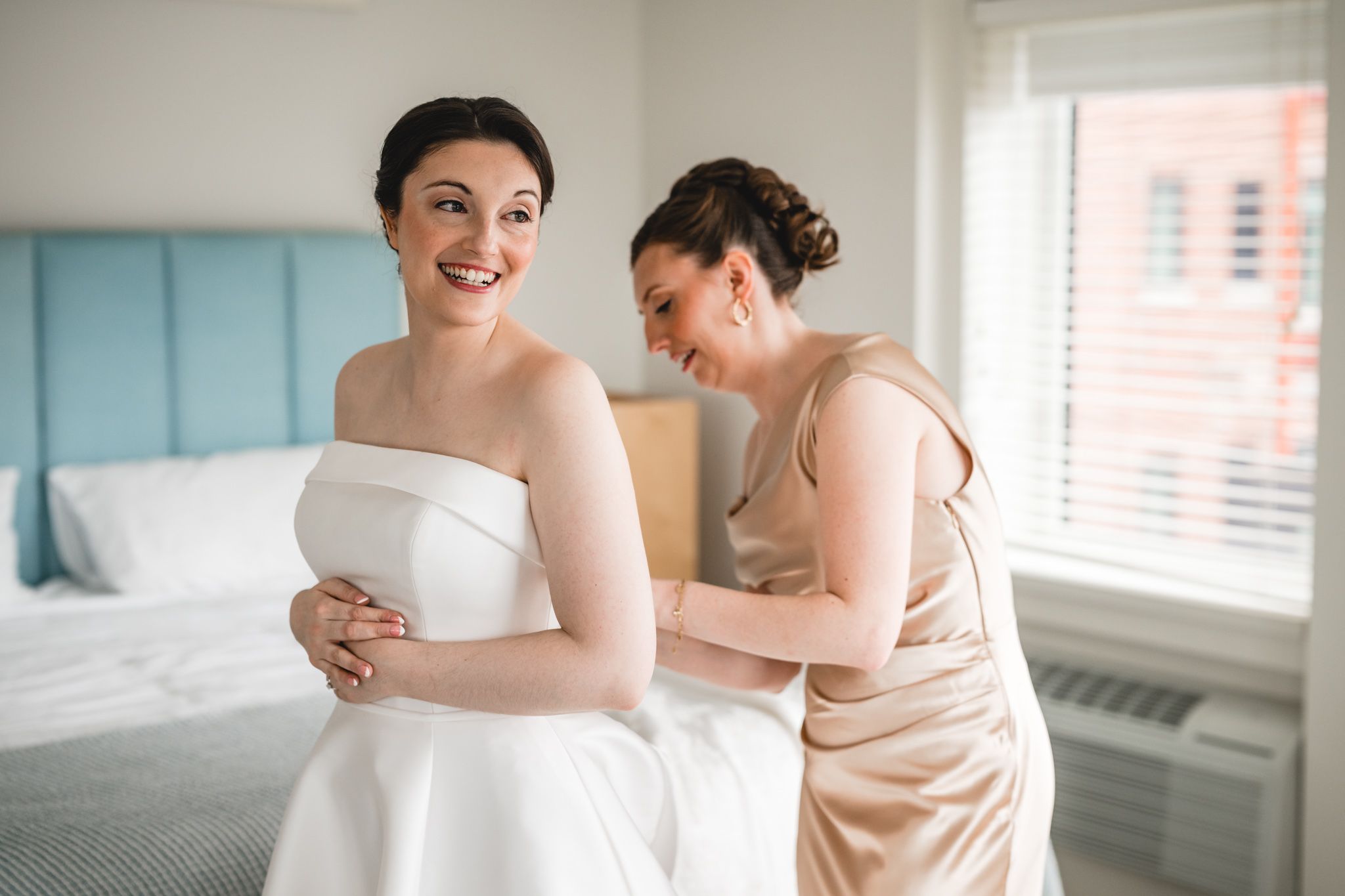Bridesmaid helping bride with white strapless wedding dress in bright hotel room.