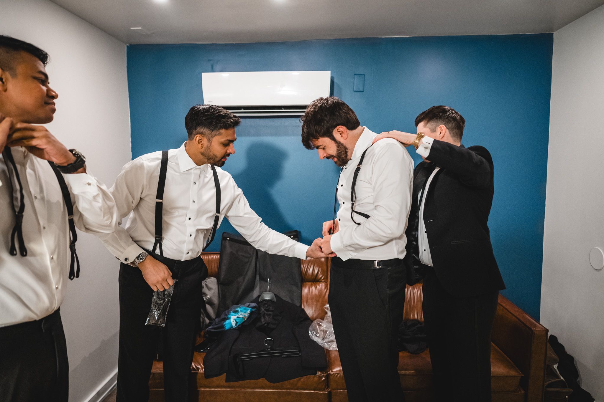 Groomsmen in white shirts and suspenders helping each other get dressed before a wedding ceremony.