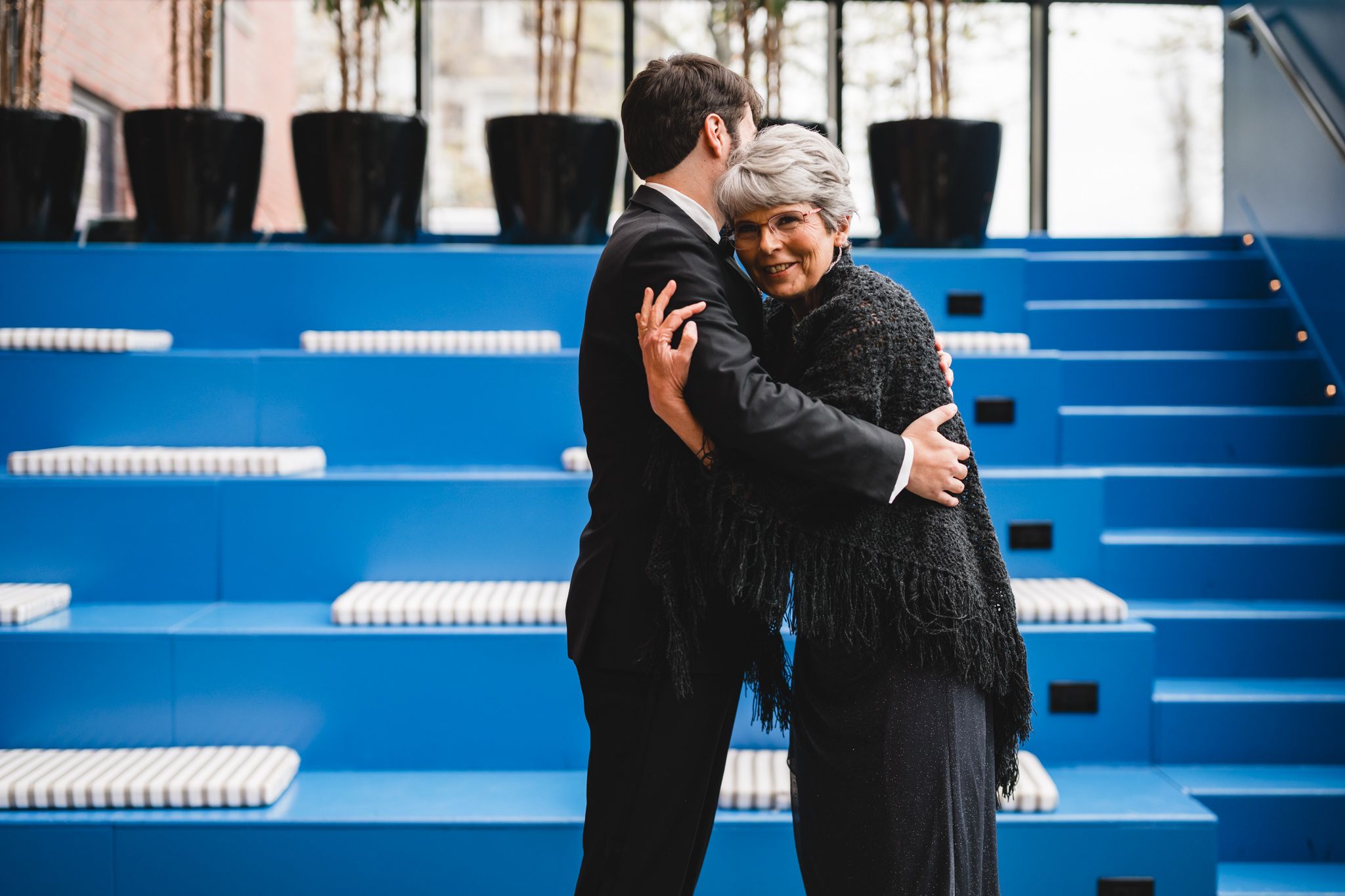 Groom in black suit sharing a warm embrace with an older woman near blue stadium seating.