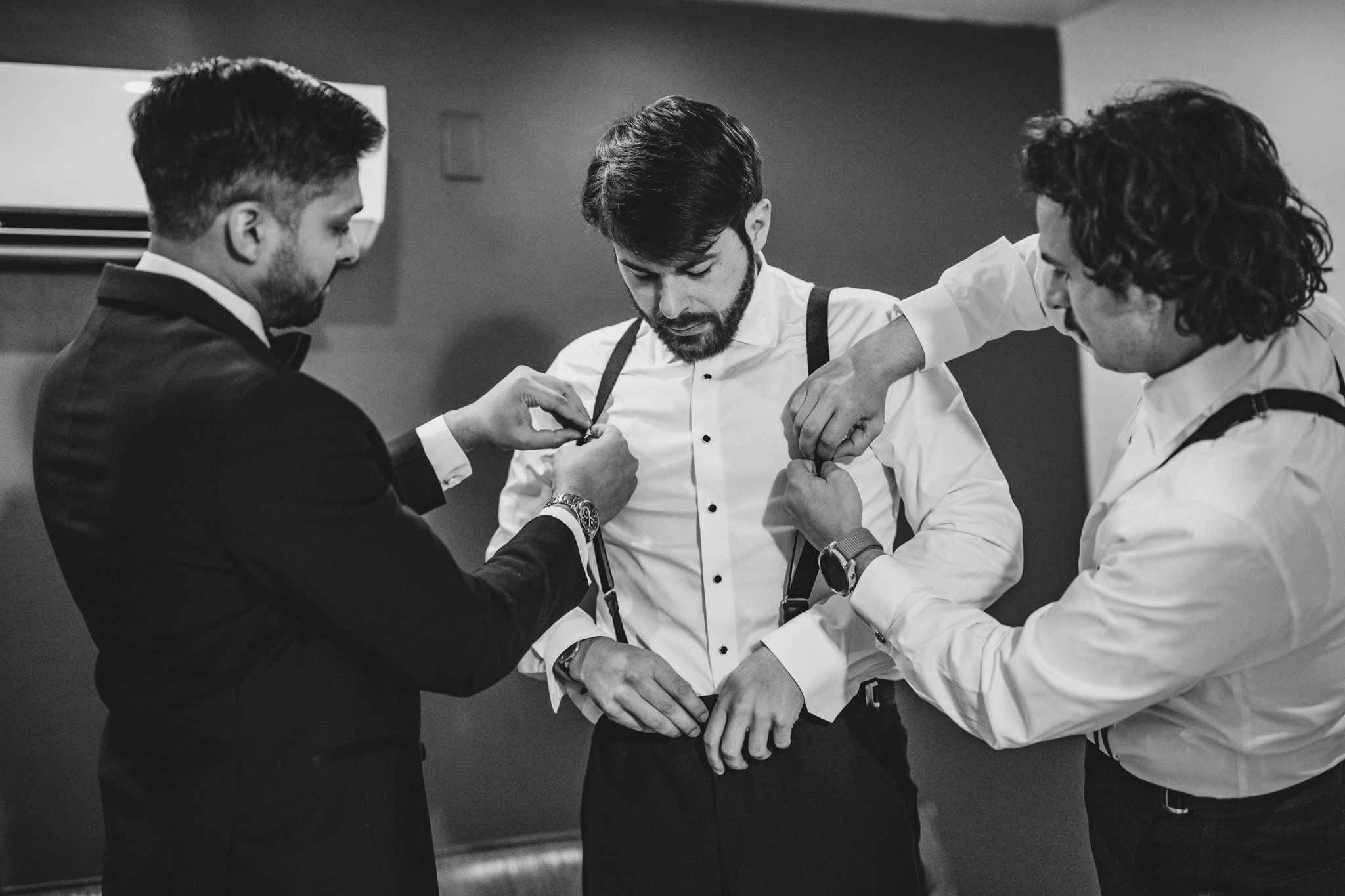 Groom getting dressed with help from groomsmen adjusting suspenders in black and white photo.