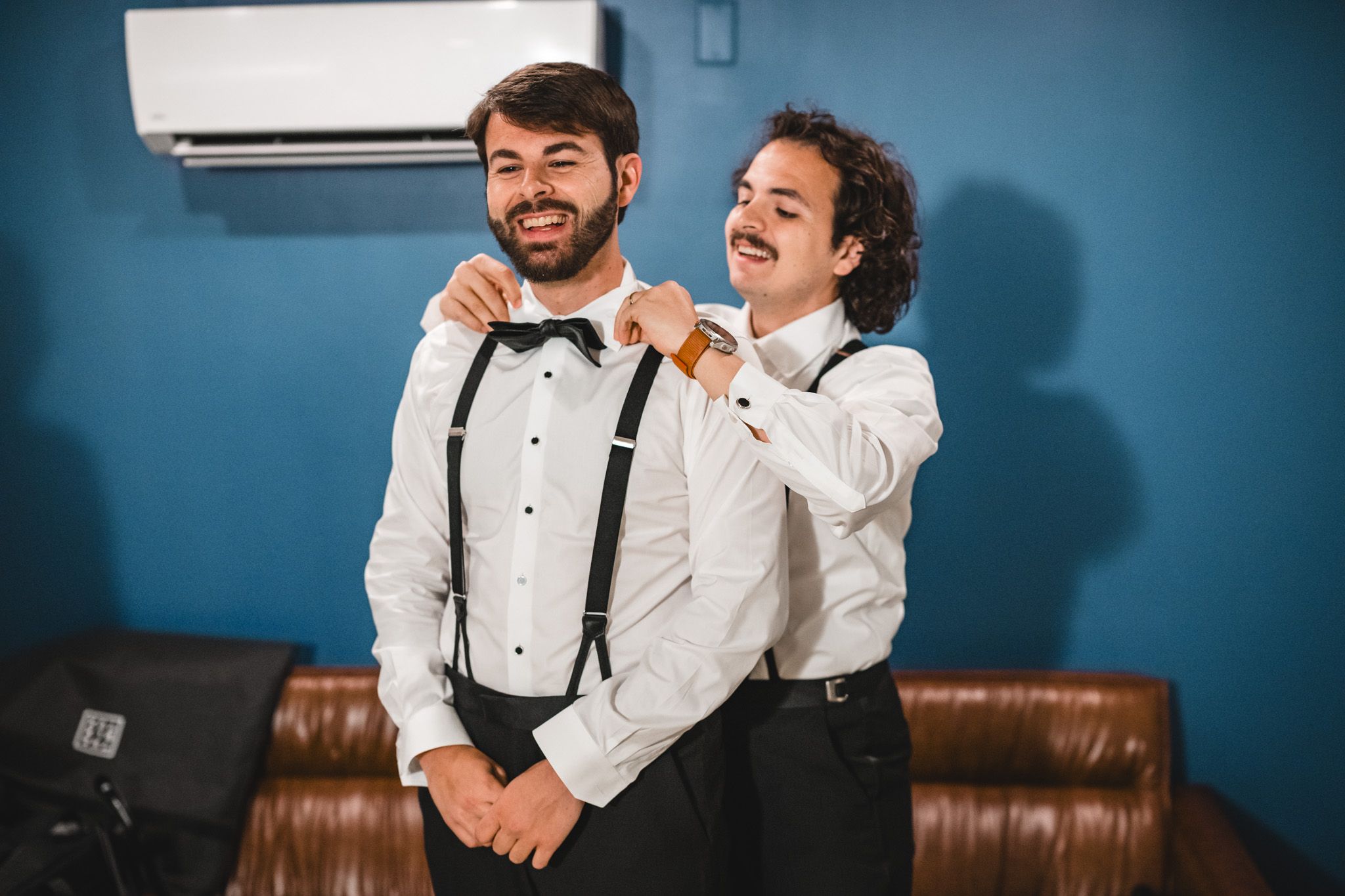 Two groomsmen in tuxedo shirts and suspenders laughing while adjusting bow tie before wedding.
