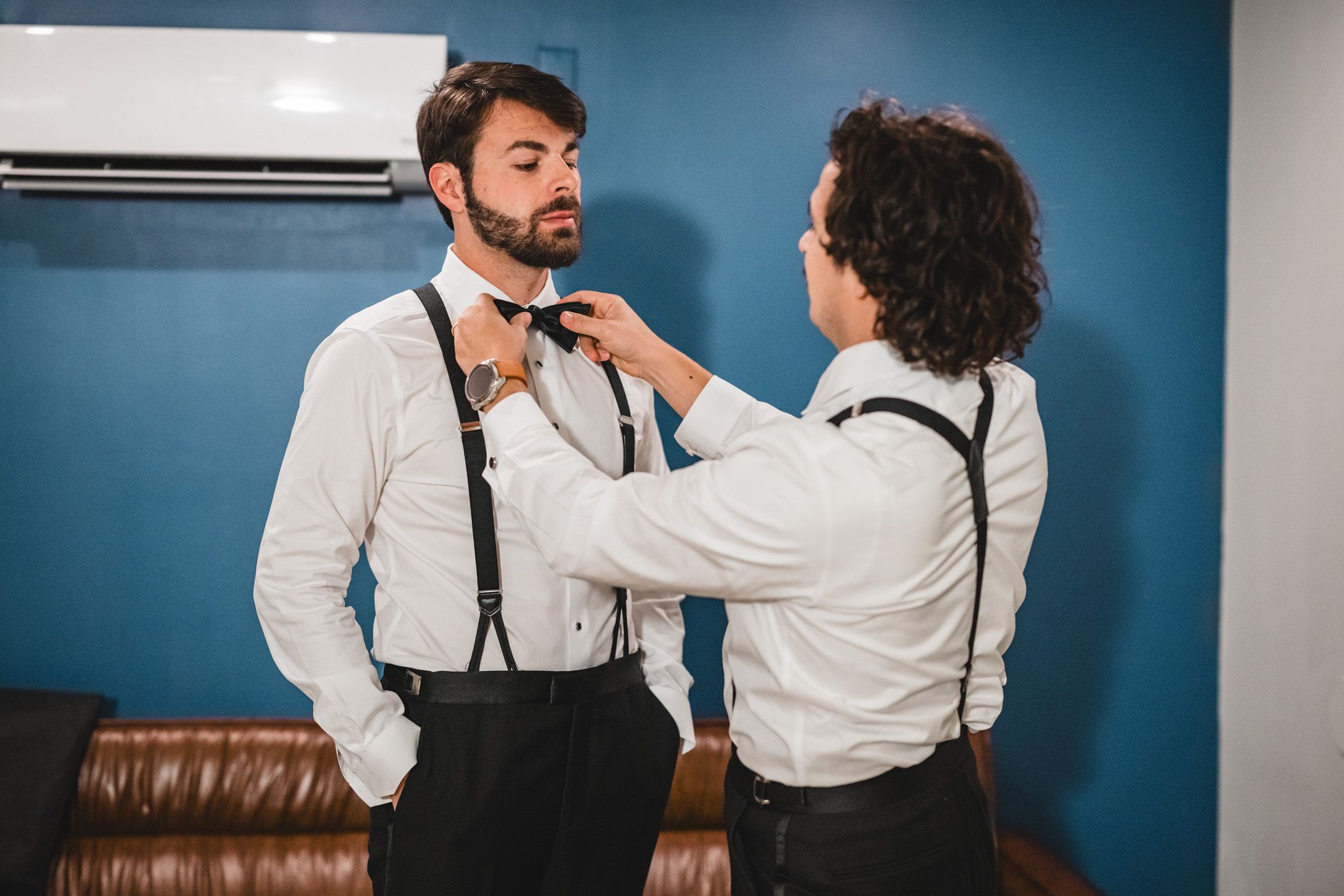 Groomsman helping groom adjust bow tie while wearing white shirts and black suspenders before wedding.