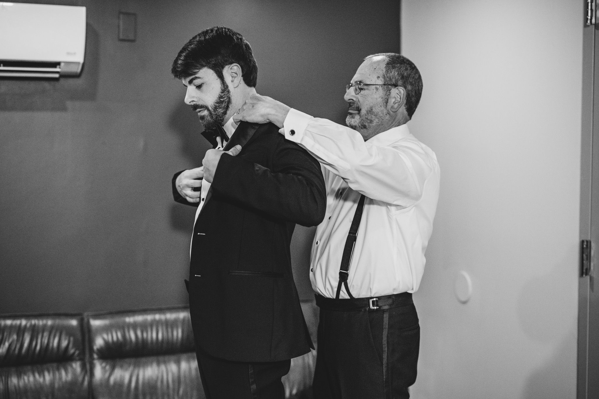 Black and white photo of a groom being helped with his suit jacket by an older man before a wedding.