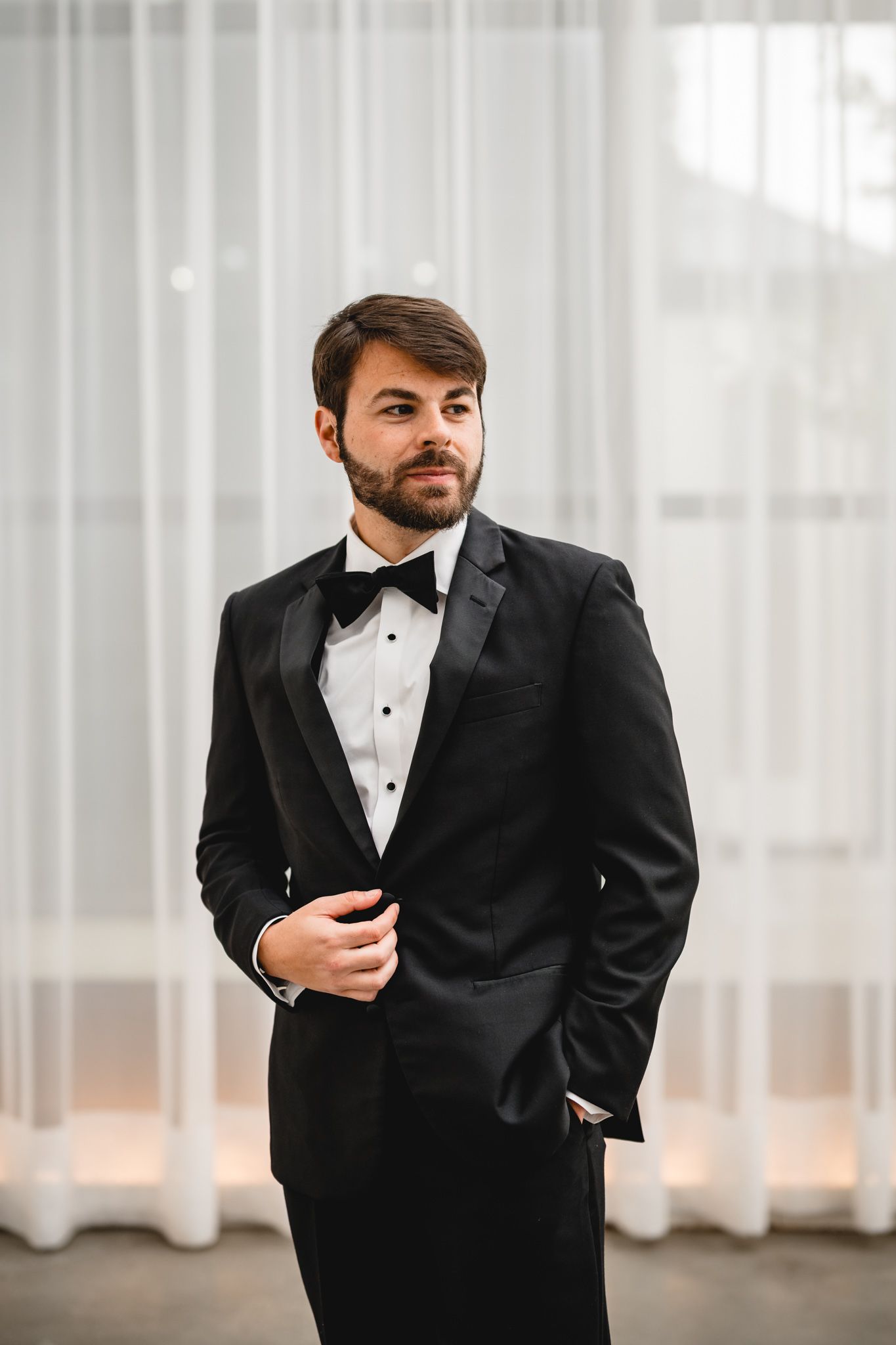 Handsome groom in black tuxedo and bow tie standing confidently in front of sheer white curtains.