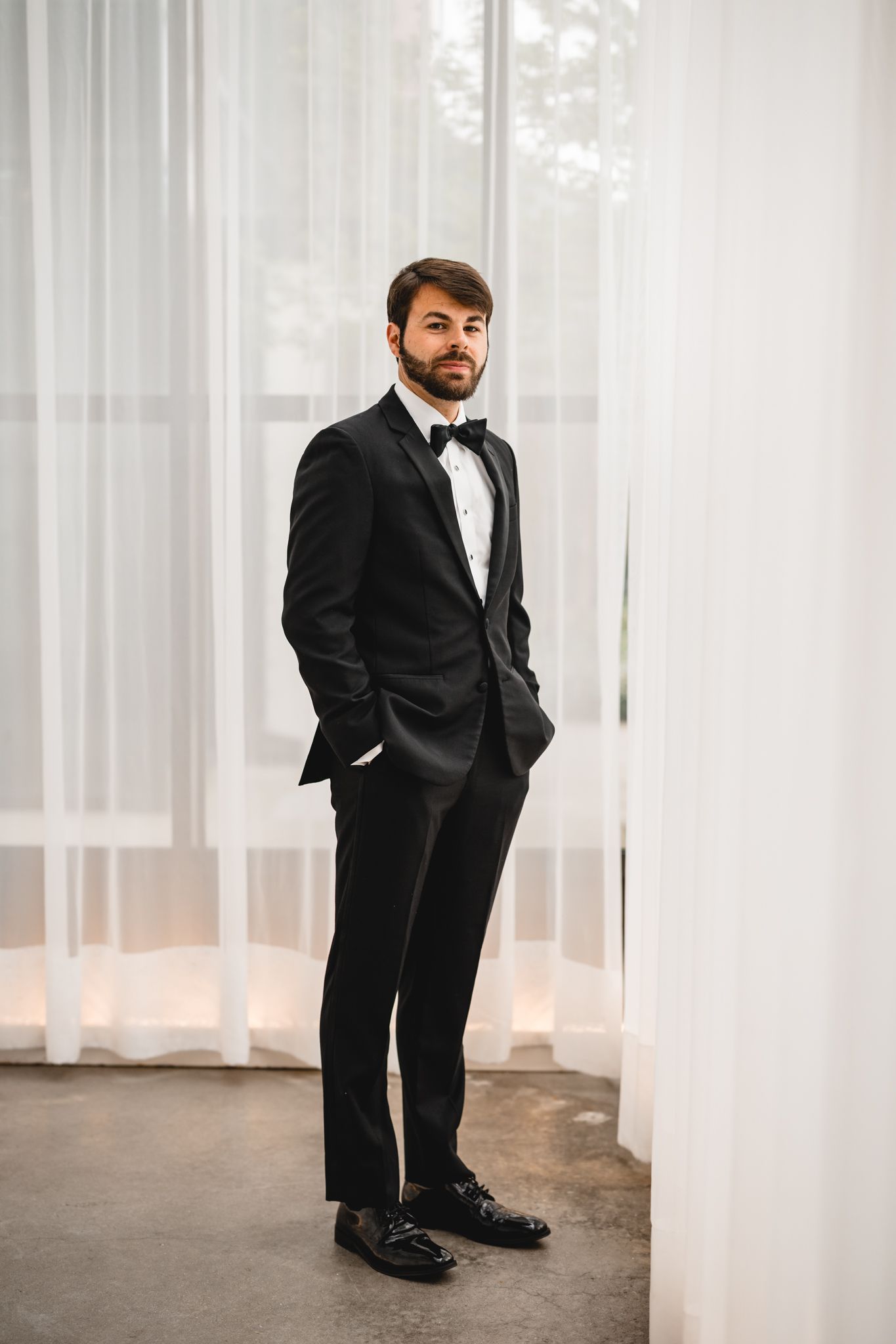 A groom in a black tuxedo with bow tie stands confidently near sheer white curtains.