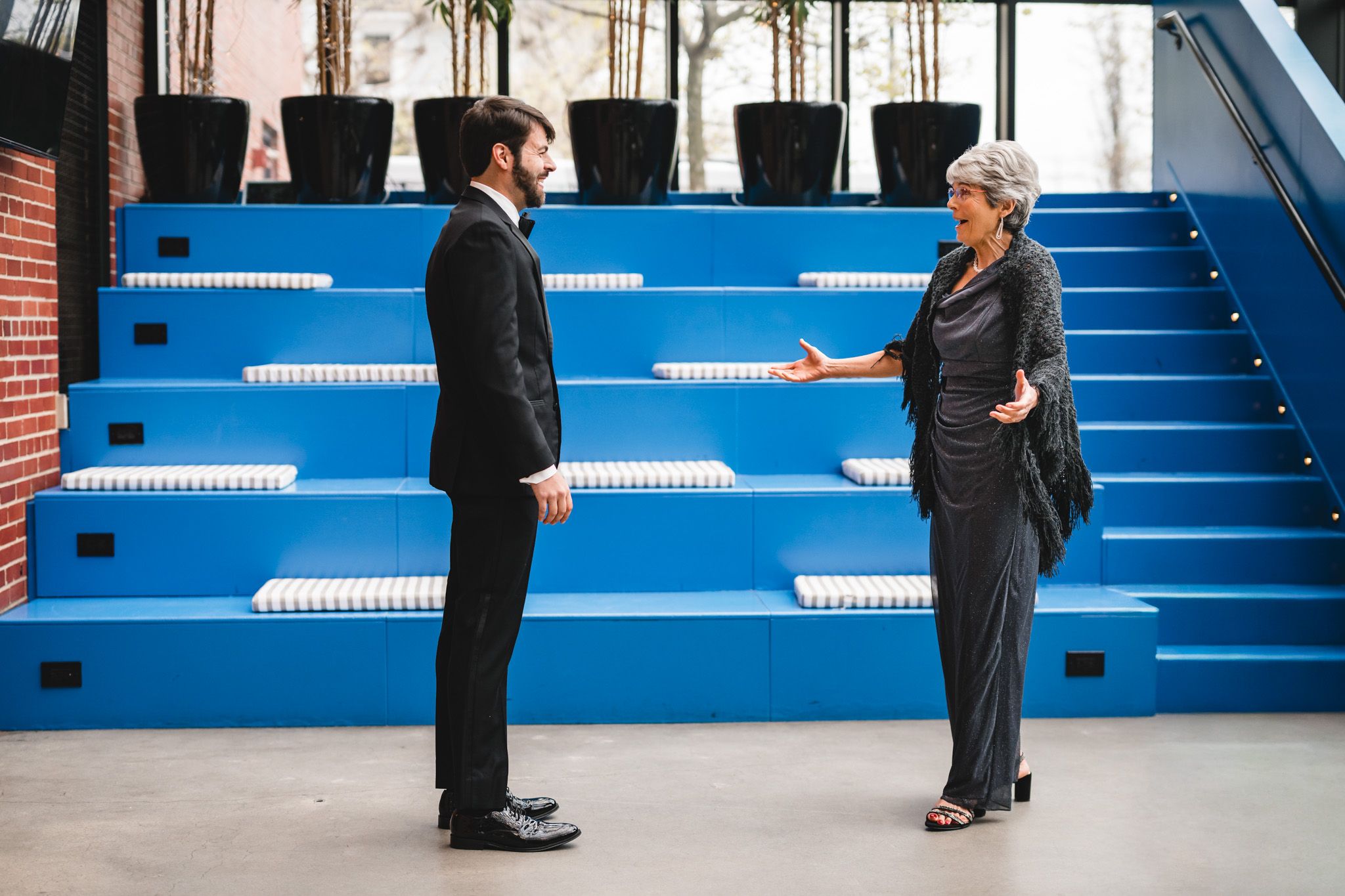 Man in black suit converses with woman in gray outfit near blue stairs in a modern venue.