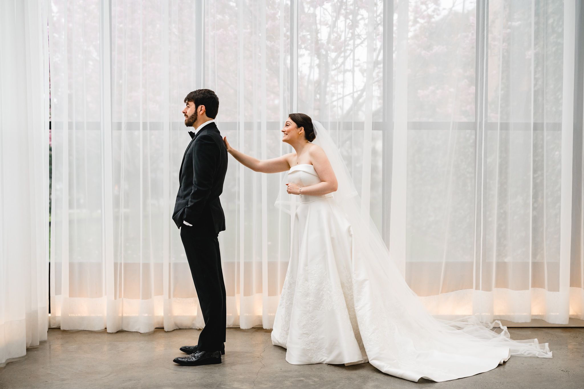 Bride in white gown touches groom's shoulder during first look moment before sheer curtain backdrop.