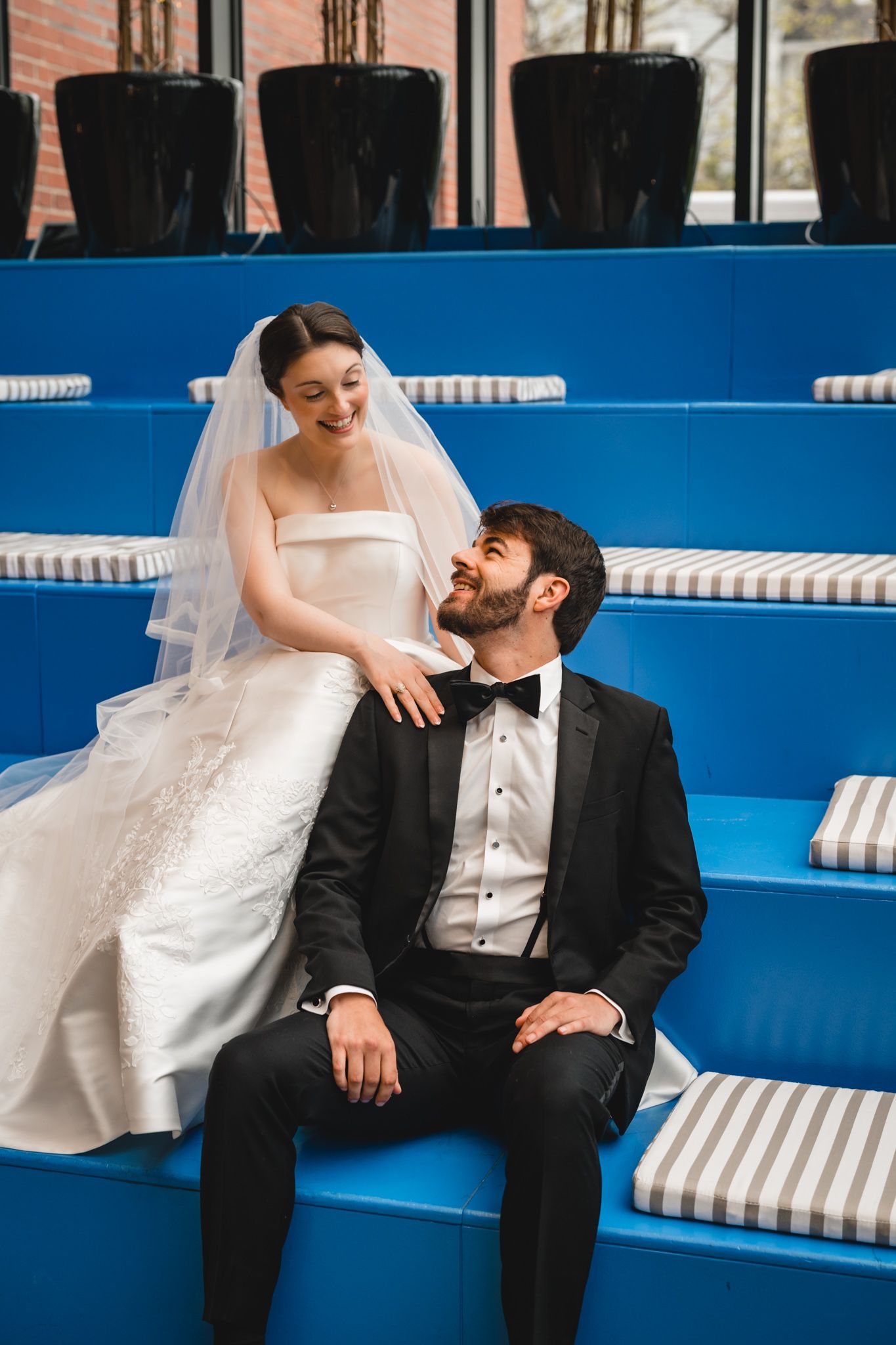 Smiling bride in white gown and veil sitting with tuxedo-clad groom on blue stadium-style seating.