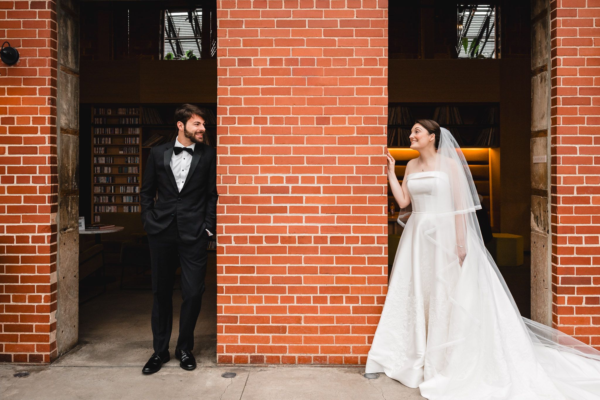 Bride in white gown and groom in tuxedo stand apart by a red brick wall, smiling at each other.