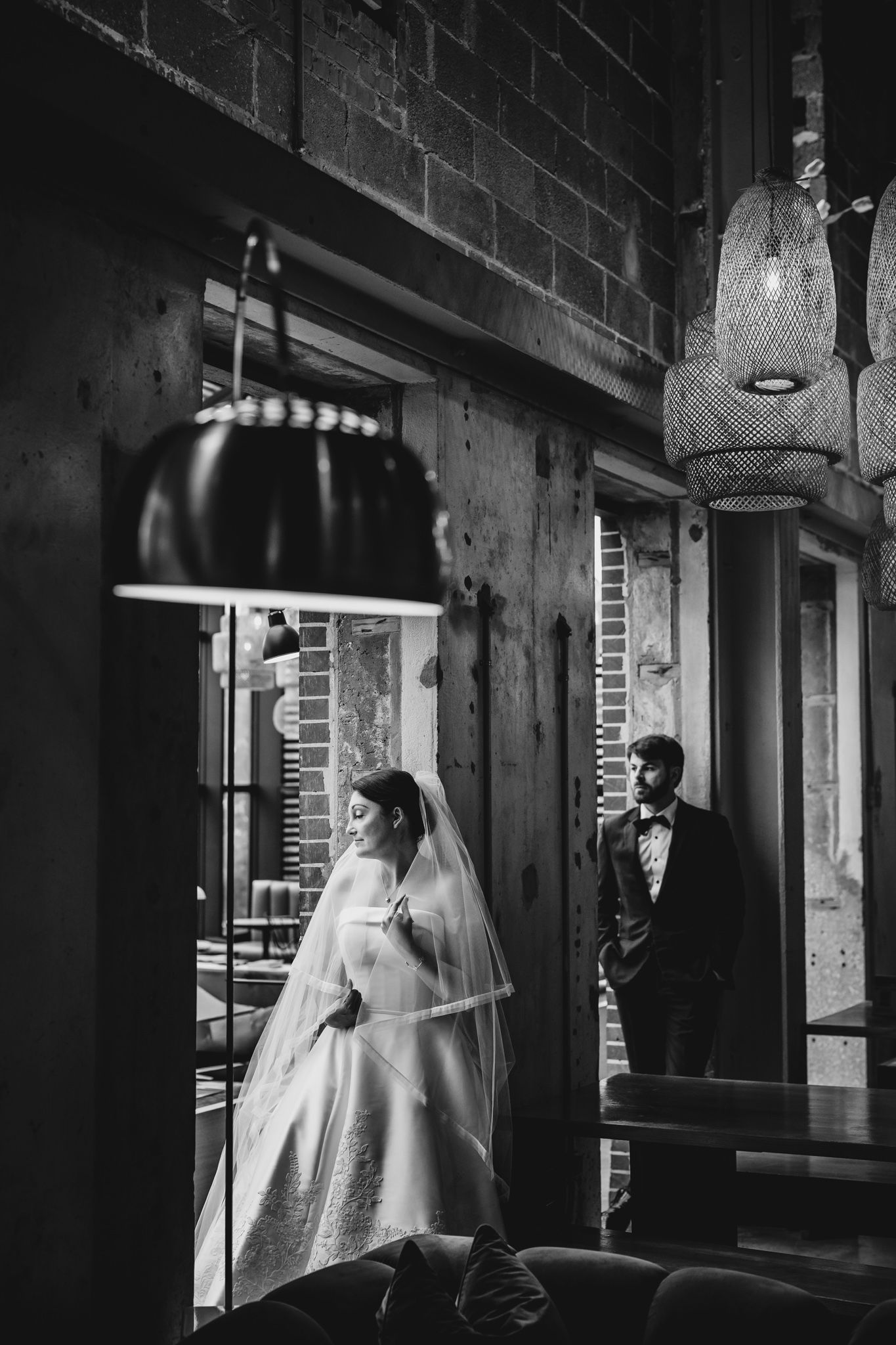 Black and white photo of bride in wedding gown and groom in tuxedo in rustic industrial venue with hanging lights.