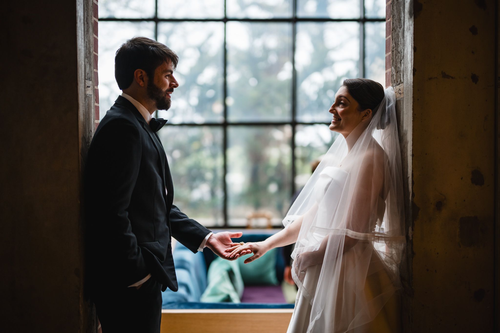 Bride and groom hold hands facing each other in romantic window light during their wedding day portrait.