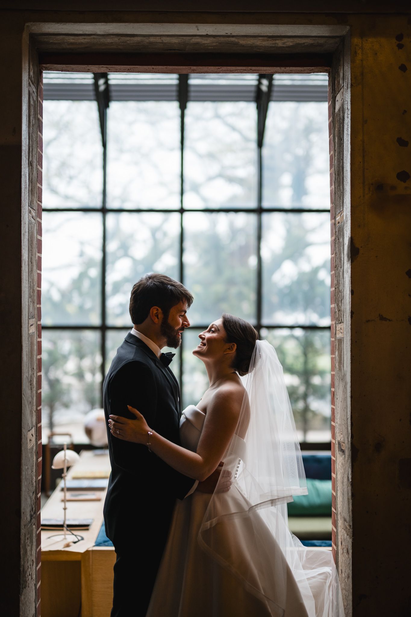 Bride and groom embracing and smiling at each other framed by a large rustic window.