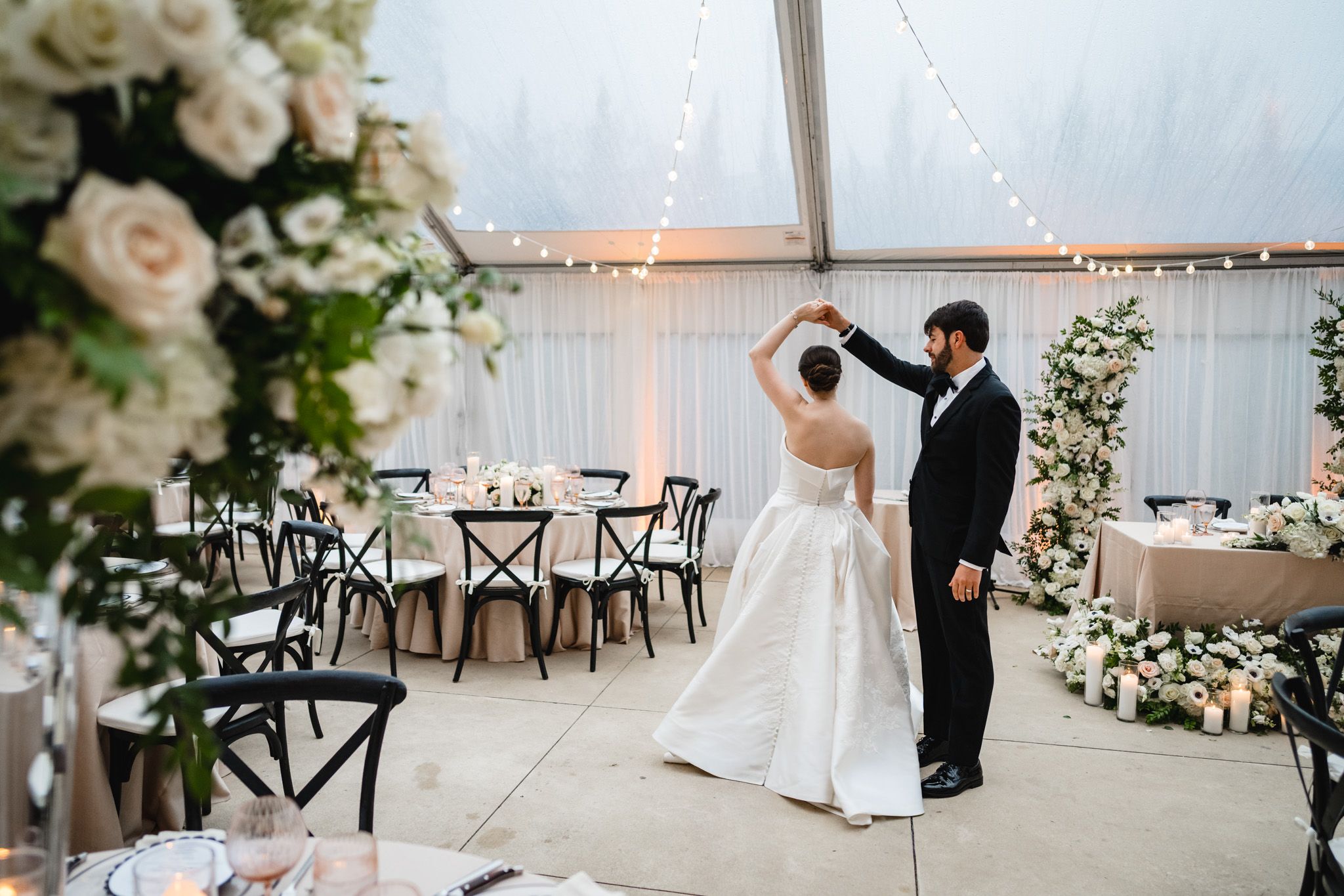 Bride and groom share first dance in elegant white tent wedding reception with floral decor.