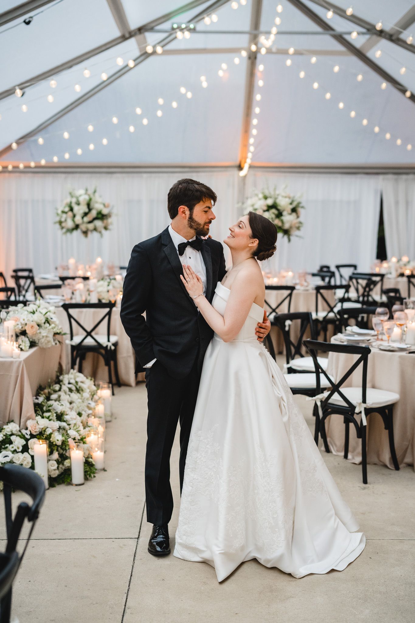 Bride and groom share a romantic moment in an elegant tented reception venue with string lights and floral arrangements.