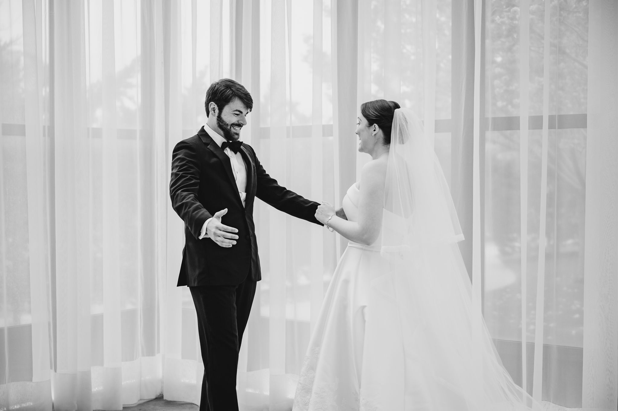 Black and white first look moment as groom in tuxedo reaches for bride in white gown and veil.