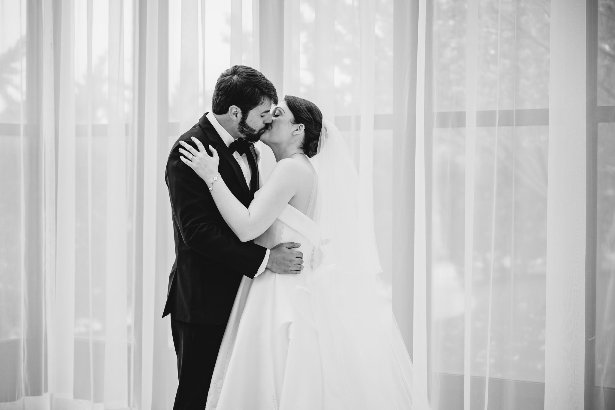 Bride and groom share a romantic kiss in black and white wedding photo near sheer curtains.