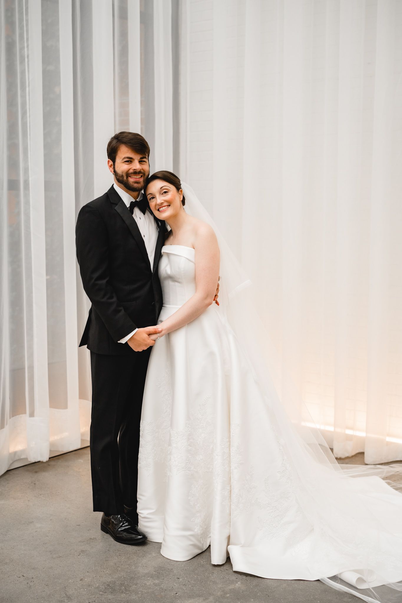 Smiling bride and groom posing together in elegant wedding attire in front of white draped backdrop.