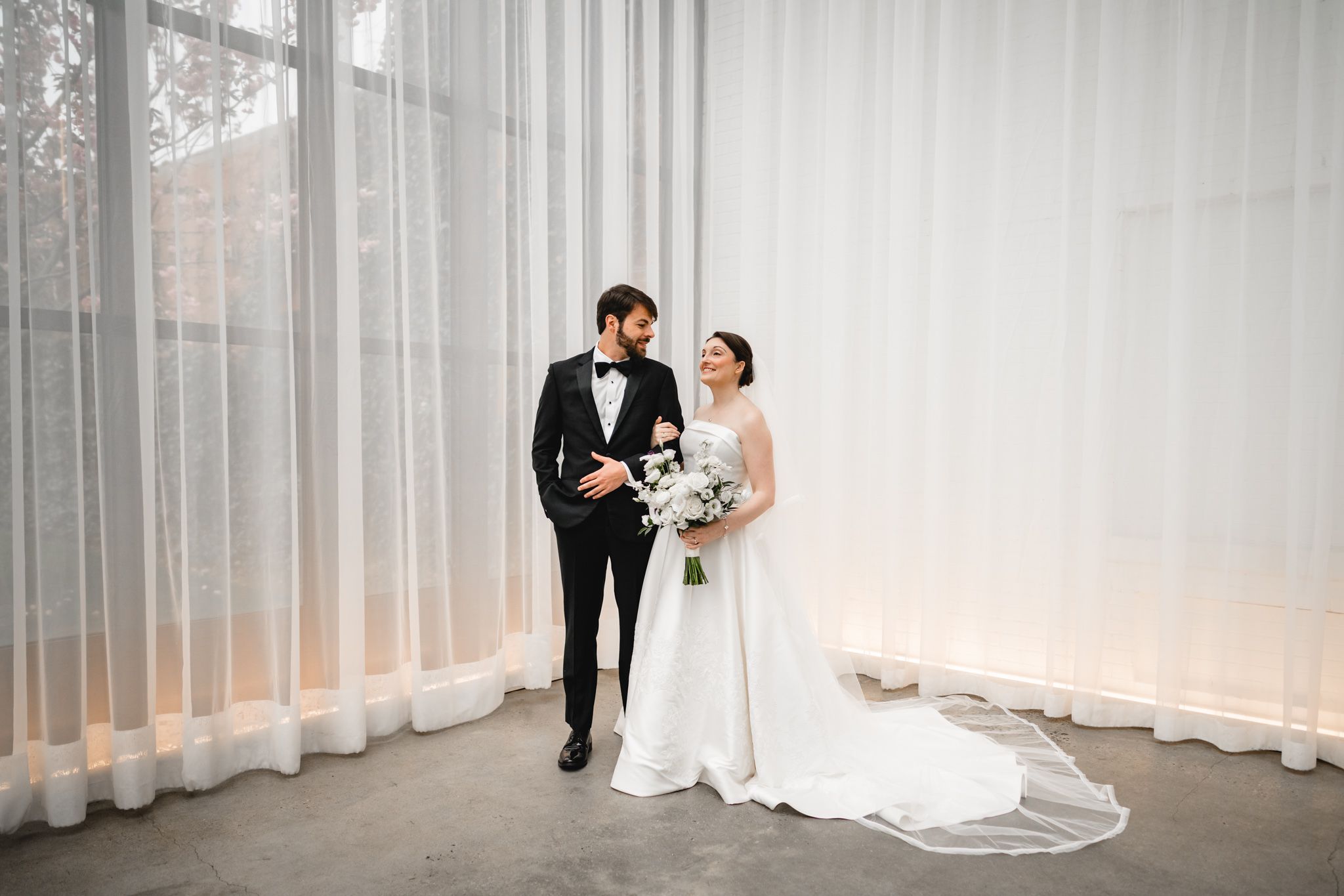 Bride and groom in elegant black tuxedo and white wedding gown pose before sheer white curtains.