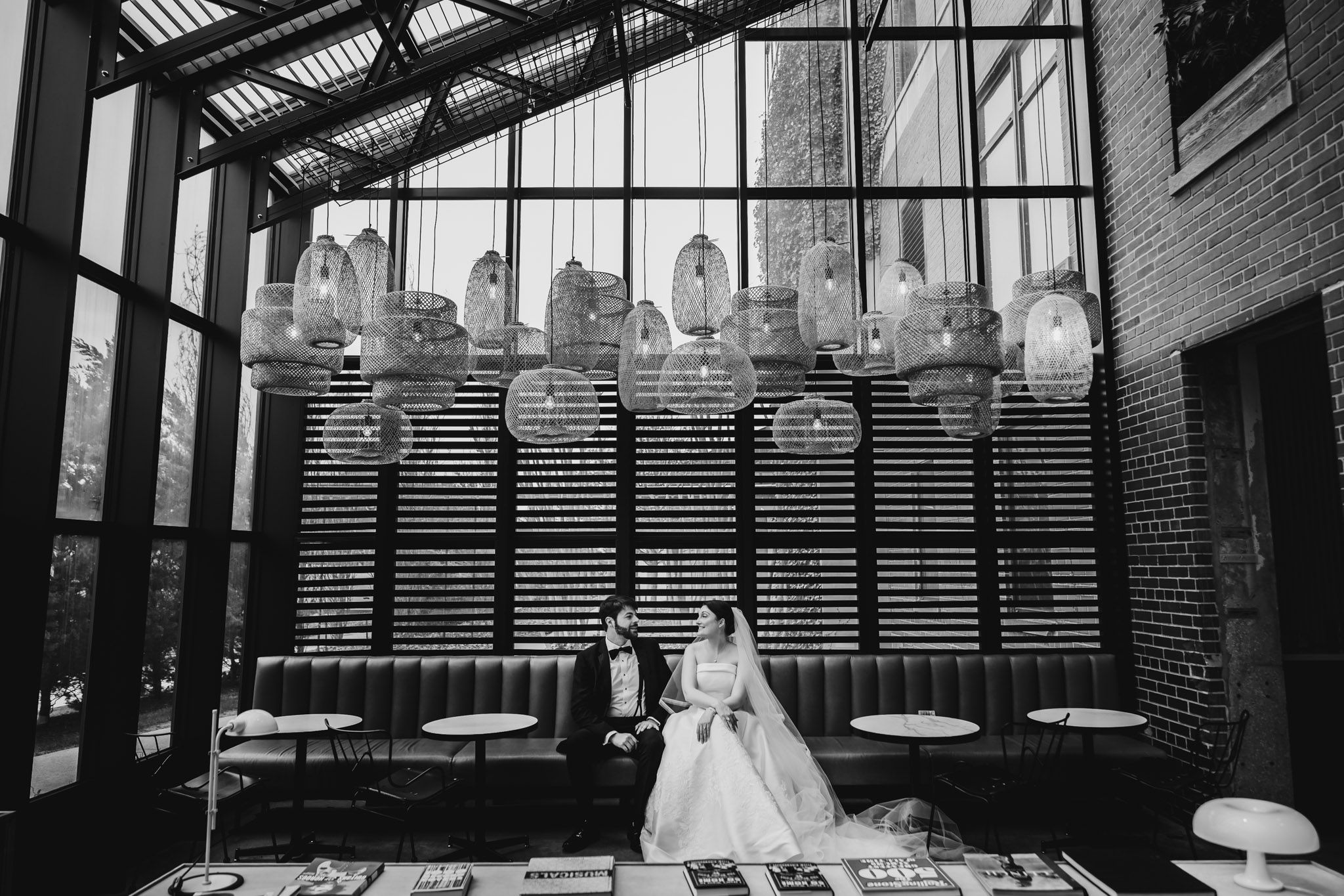 Bride and groom sit together in elegant black and white photo inside a stylish venue with hanging pendant lights.